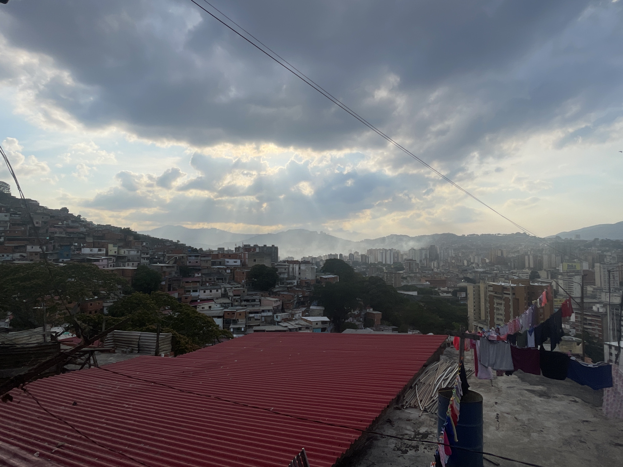 The image shows an urban view of Caracas, Venezuela, under a cloudy sky with sunlight breaking through. The foreground features a corrugated red roof and a rooftop with clothes hanging on a clothesline. In the midground, a densely packed hillside settlement sprawls across the landscape. The background shows a cityscape with mountains in the distance. There are no people visible.