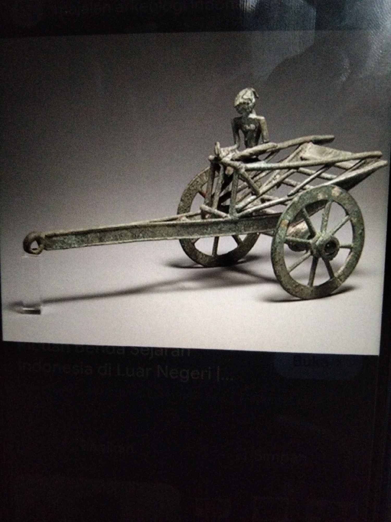 A bronze miniature chariot with a figure is displayed against a plain, light grey background. The chariot has two spoked wheels, a long rectangular body, and a shaft extending forward with a hook at the end. A small, crudely formed figure of a person is seated at the front of the chariot, holding onto the structure. The chariot appears old and weathered, with a green patina on the bronze. There is a shadow cast by the chariot on the surface it rests on. Text is visible at the bottom of the image, indicating it is part of a collection of historical artifacts, possibly related to Indonesia. The specific location within Kualaraja, Indonesia, is not discernible from the image.