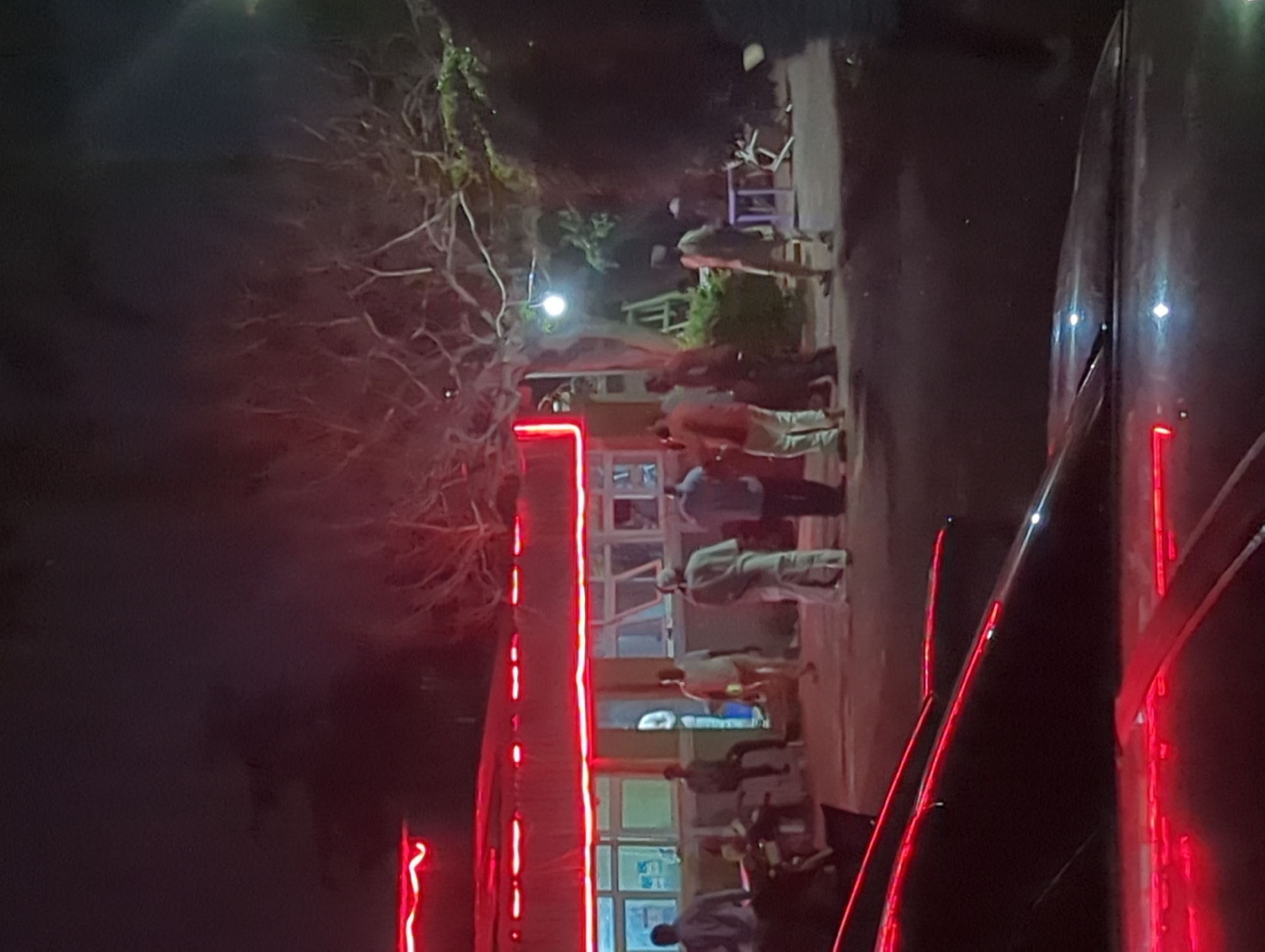 In Narayi, Nigeria, at night, several individuals are gathered outside a building illuminated by bright red neon lights. The building appears to be a commercial establishment with multiple windows. Some people are standing in groups, while others are seated. One person is seen walking towards the building. In the foreground, the hood of a dark-colored car with red neon reflections is visible. The environment is dark, with the red lights creating a prominent visual element. The background shows trees and some outdoor furniture, suggesting a public or commercial space.