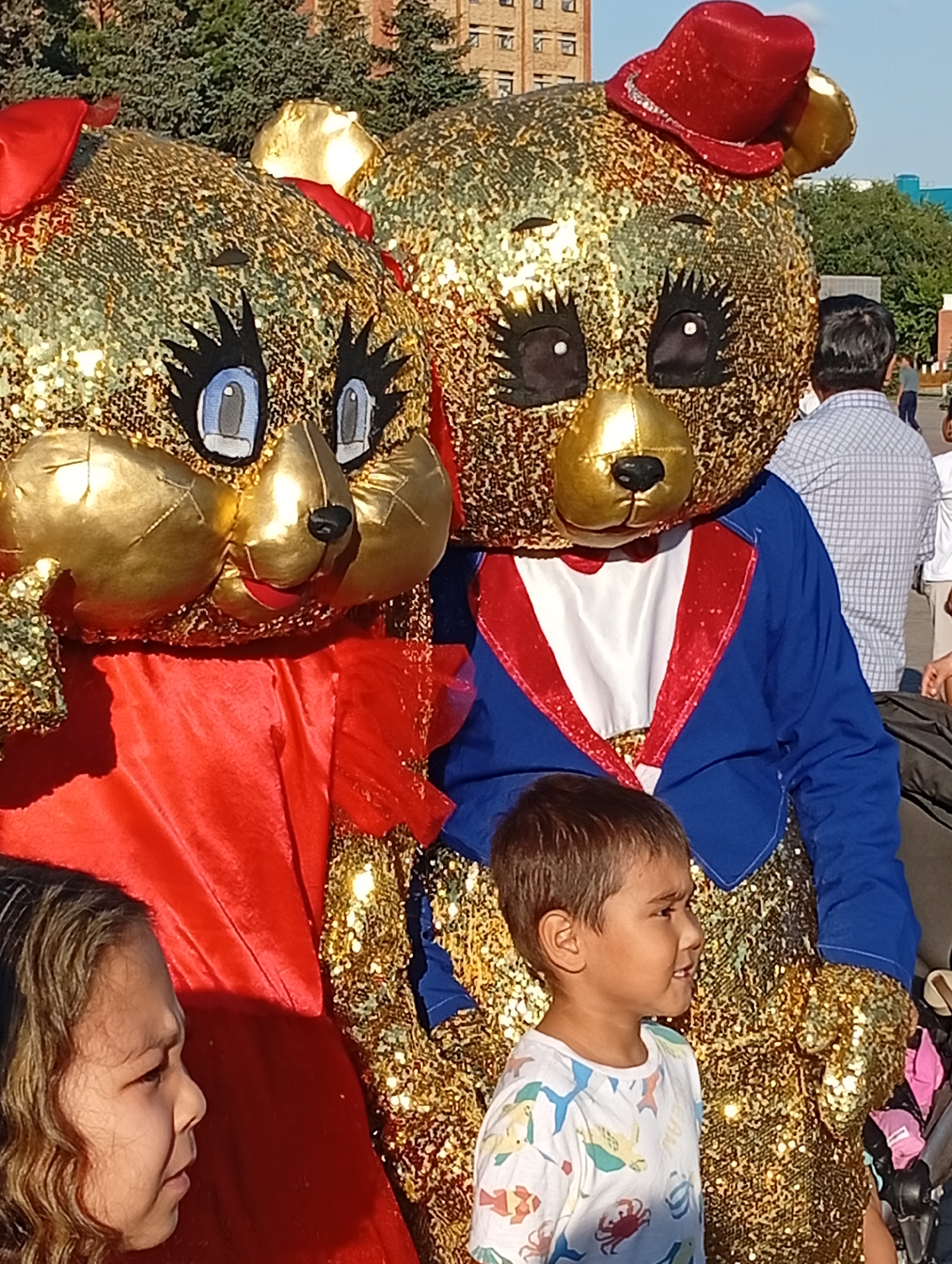 The image depicts an outdoor scene with two people in bear costumes and two children in the foreground, possibly taken in Kostanay, Kazakhstan, given the buildings and trees visible in the background.
The two bear costumes are large and covered in gold sequins. The bear on the left has a red bow on its head and is wearing a red dress. The bear on the right wears a red top hat with a silver band and a blue jacket with red trim over a white shirt and red bowtie. Both costumes have large, cartoonish eyes with prominent eyelashes and gold snout and ear details.
In the foreground, a young boy is looking to his right, wearing a white t-shirt with a colorful pattern of sea creatures. To his left, the side profile of a young girl with curly brown hair is visible, her gaze also directed to her right.
The background shows trees and buildings, suggesting an urban environment. The lighting suggests it is daytime. The overall scene appears to be a festive or public gathering.
