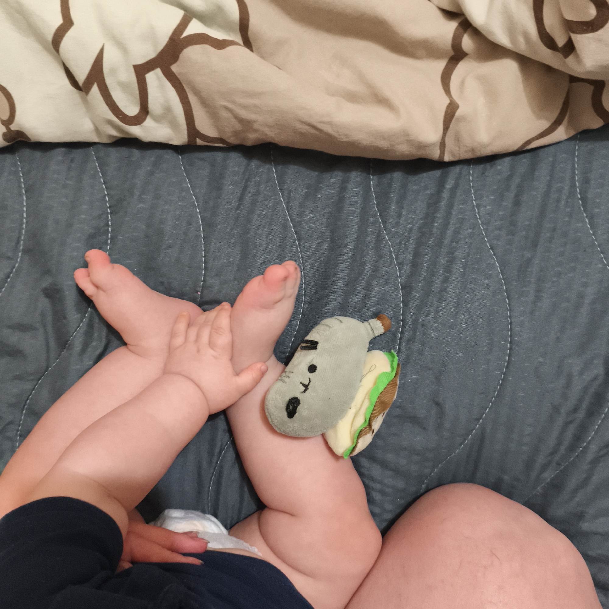The image shows a baby lying on a gray quilted blanket with a patterned beige blanket at the top edge. The baby is wearing a navy blue shirt and a diaper. Their legs are crossed, and their hands are touching their feet. A gray stuffed toy shaped like a cat inside a sandwich is resting on the baby's thigh. The setting appears to be indoors, likely a home. There is no information to determine the location is Chaikovsky, Russia.