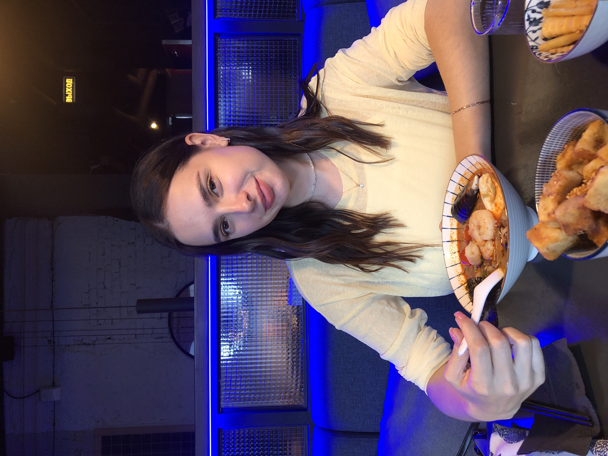 A young woman with long, dark hair is seated at a table in what appears to be a restaurant. She is wearing a light yellow, long-sleeved shirt and a delicate necklace. The woman is holding a spoon and is about to eat from a bowl of soup, which contains shrimp, mussels, and other ingredients. In front of her, there are two other bowls, one filled with fried food and another with what looks like pickled vegetables. The background is dimly lit with blue neon lighting. There is a sign on the wall that says "ВЫХОД" (Exit). The woman's expression is pleasant, and she is looking towards the camera. The scene suggests a casual dining experience. The location of this restaurant is in Izhevsk, Russia.