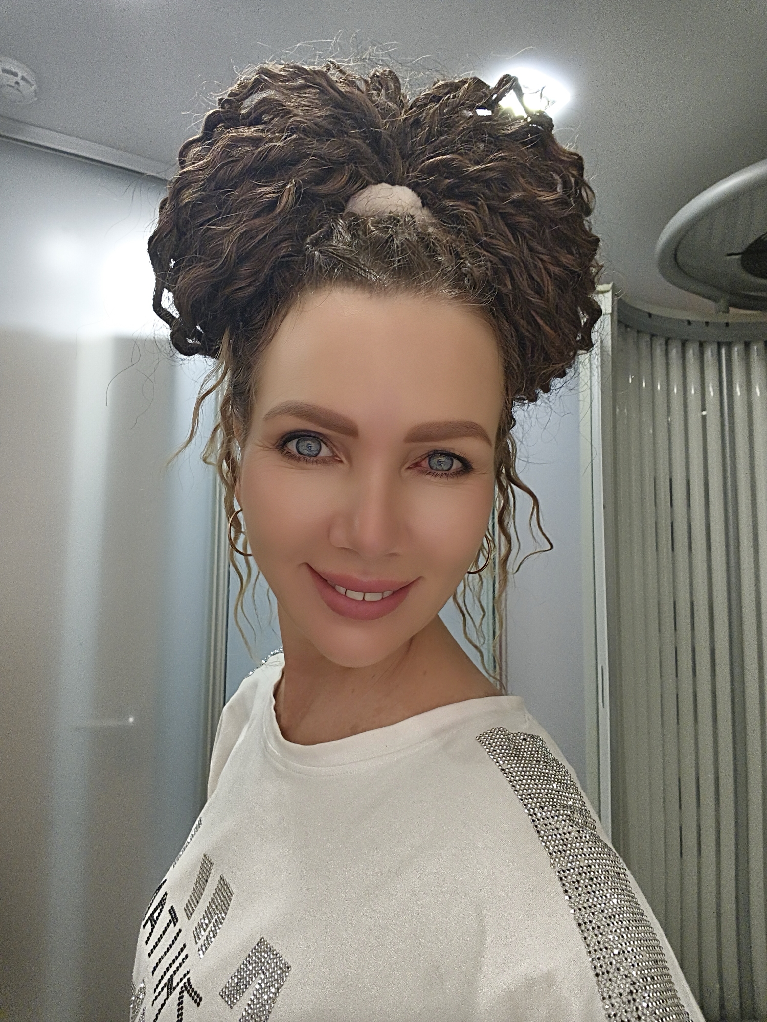 A woman with curly brown hair styled in a bun and loose tendrils around her face is pictured in a close-up, eye-level shot. She has blue eyes, prominent cheekbones, and is smiling with her lips slightly parted, revealing her teeth. She is wearing a white t-shirt with what appears to be sparkling embellishments on the shoulders and chest. The background is blurred, but suggests an indoor setting with vertical blinds and possibly a mirrored or metallic surface. The lighting is bright, with a spotlight visible in the upper right portion of the frame. The scene does not provide enough information to determine a specific city or location.
