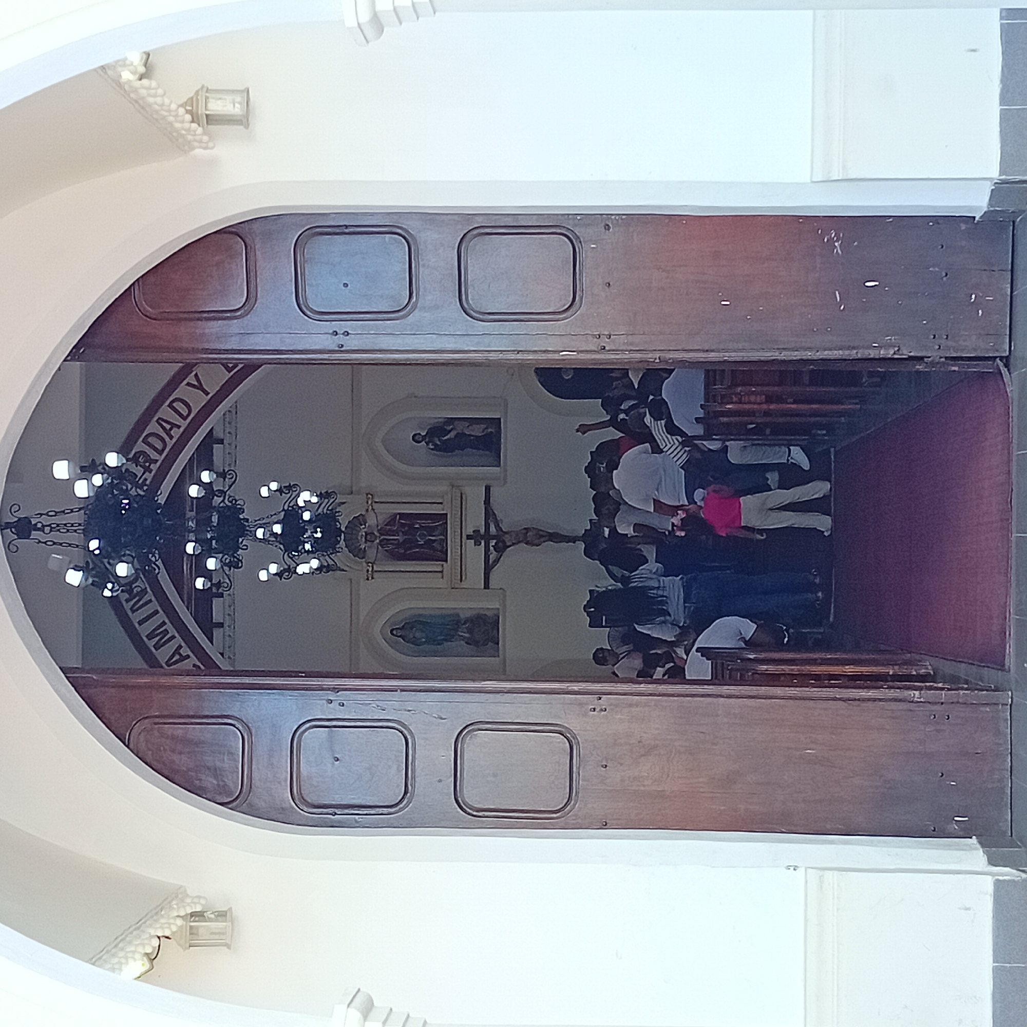 The photograph captures a scene from inside a church. The view is through a large, arched doorway with ornate wooden doors open to reveal the interior.
Inside the church, there's a chandelier hanging from the ceiling. The interior architecture features religious paintings and a crucifix mounted on the wall. A group of people are seated in pews. A maroon carpet runs down the aisle between the pews.
The arched doorway is part of a white structure, which forms the entrance to the church. Small lamps are mounted on the wall on either side of the arch.