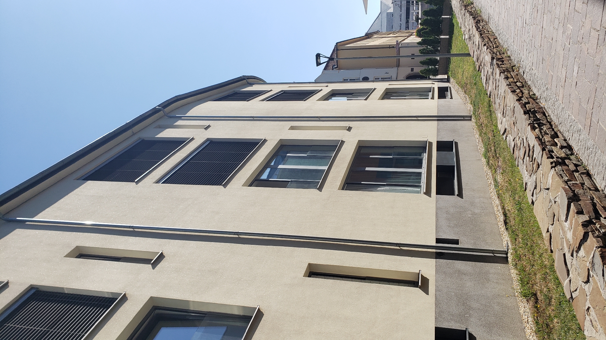 The image shows a modern apartment building with a light beige facade, located on a sunny day in Prešov, Slovakia. The building features multiple floors with large windows, some of which are equipped with dark roller shutters. A metallic downspout runs vertically along the side of the building. In front of the building, there is a low, uneven stone wall bordering a grassy area. A paved walkway runs alongside the stone wall, and a lamppost stands further down the path. The sky is clear blue, indicating fair weather. In the background, other buildings and some evergreen trees are visible. The scene is devoid of any people or vehicles.