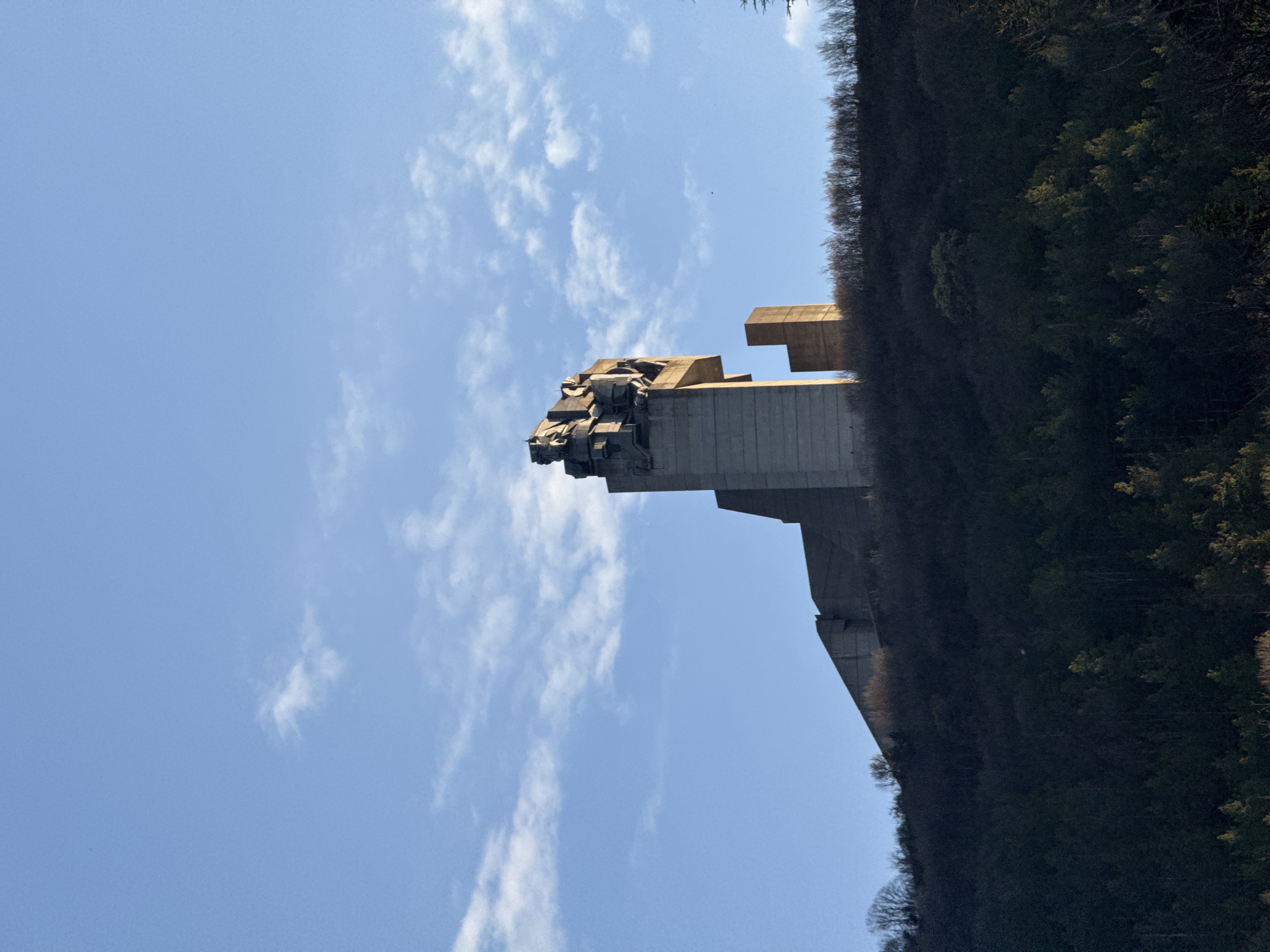 The image shows the Founders of the Bulgarian State Monument in Shumen, Bulgaria. The monument is a large concrete structure with stylized figures at the top, positioned on a hillside. The sky is blue with scattered white clouds. A heavily wooded area covers the hillside to the right of the monument.