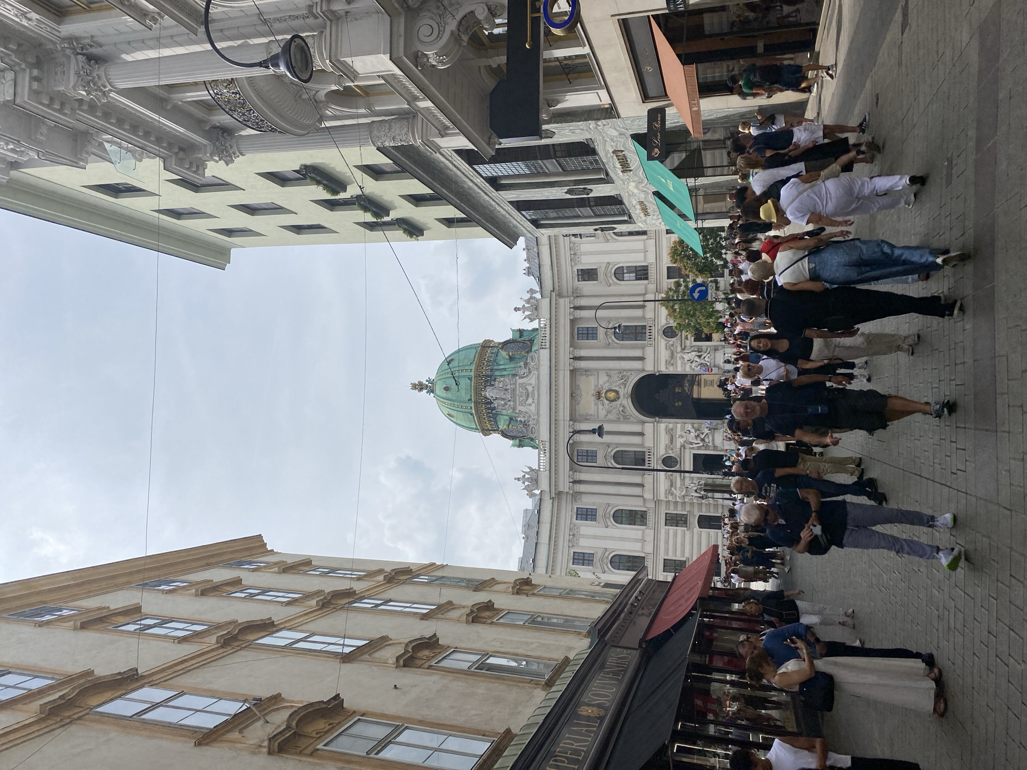The image depicts a street scene in Vienna, Austria, specifically in front of the Hofburg Palace. A pedestrian street is lined with buildings showcasing classical architecture. On the left, an ornate building with multiple windows and decorative elements houses "Imperial Souvenirs." To the right, other buildings feature elaborate facades, balconies, and shops. In the background, the prominent green dome of the Hofburg Palace is visible. A crowd of people, dressed in casual attire, are walking along the cobblestone street, some looking at their phones, others engaged in conversation. The sky is overcast with scattered clouds.