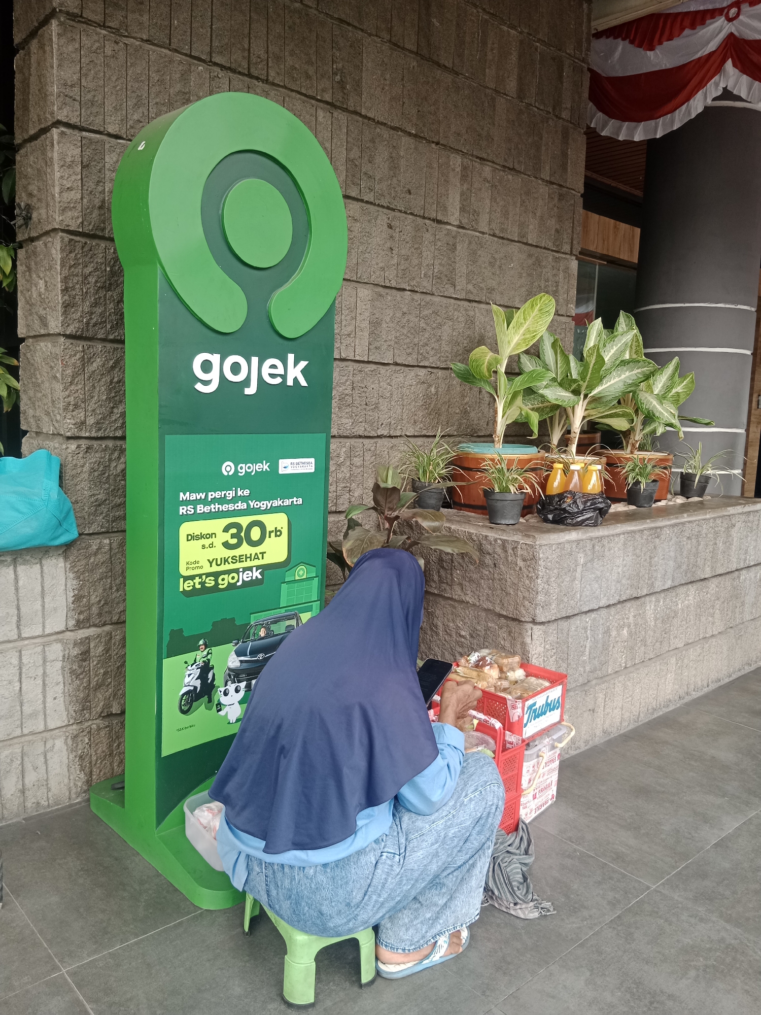 A person wearing a dark blue hijab and light blue jeans sits on a green plastic stool next to a large green Gojek advertisement pillar. The pillar displays the Gojek logo and promotional text in Indonesian. To the right of the pillar, a woman is handling items in a red plastic crate, which appears to contain food items. The Gojek pillar is positioned in front of a stone-tiled wall. In the background, several potted plants are visible on a ledge. The scene suggests an outdoor setting, likely a public area in Yogyakarta, Indonesia, given the Gojek branding and the Indonesian text on the advertisement.