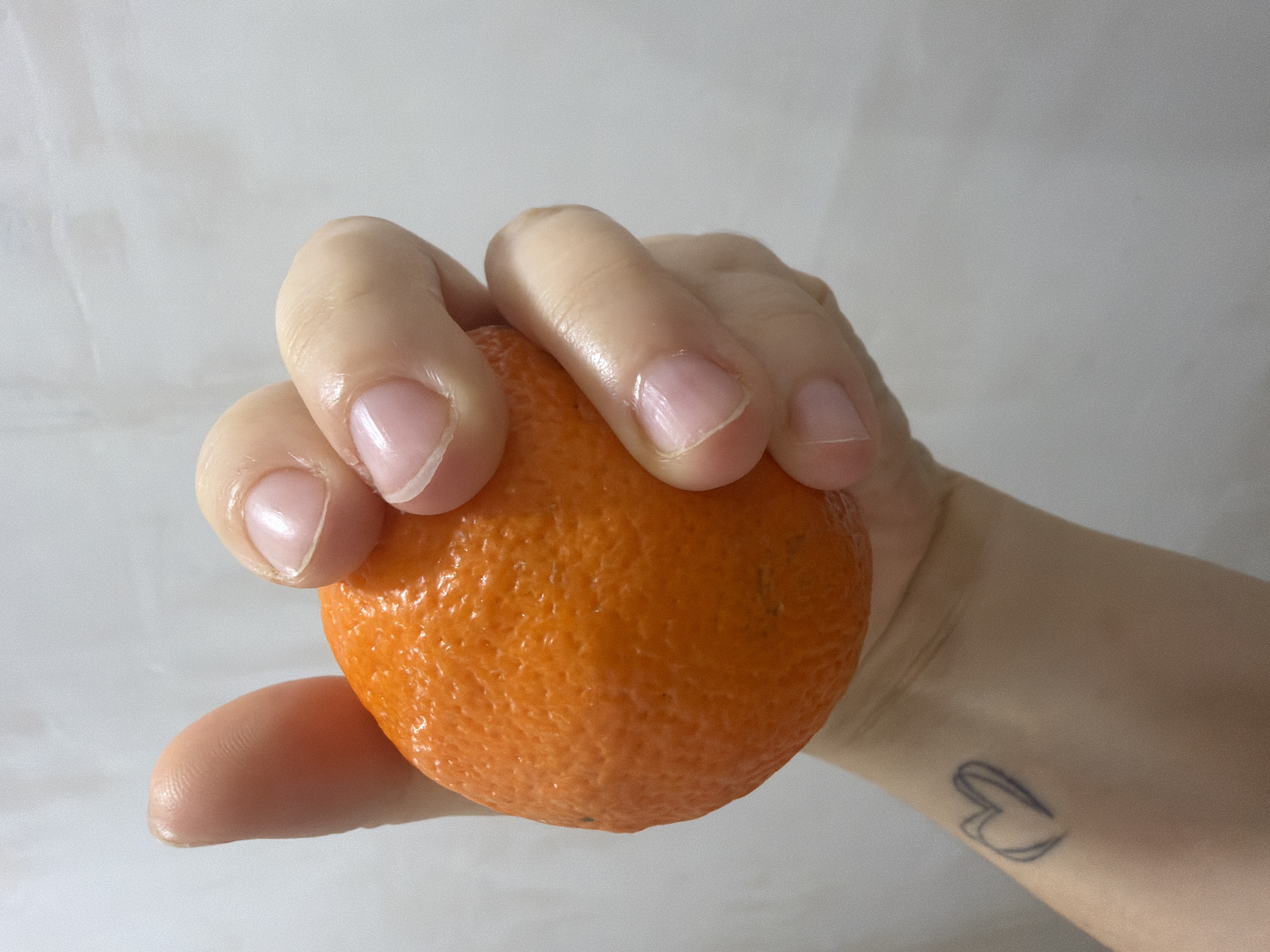 A hand is holding an orange against a light grey background. The hand appears to be light-skinned with short, well-groomed nails. A tattoo of a heart outline is visible on the wrist. The orange is textured and orange in color. The location is not discernible from the image.