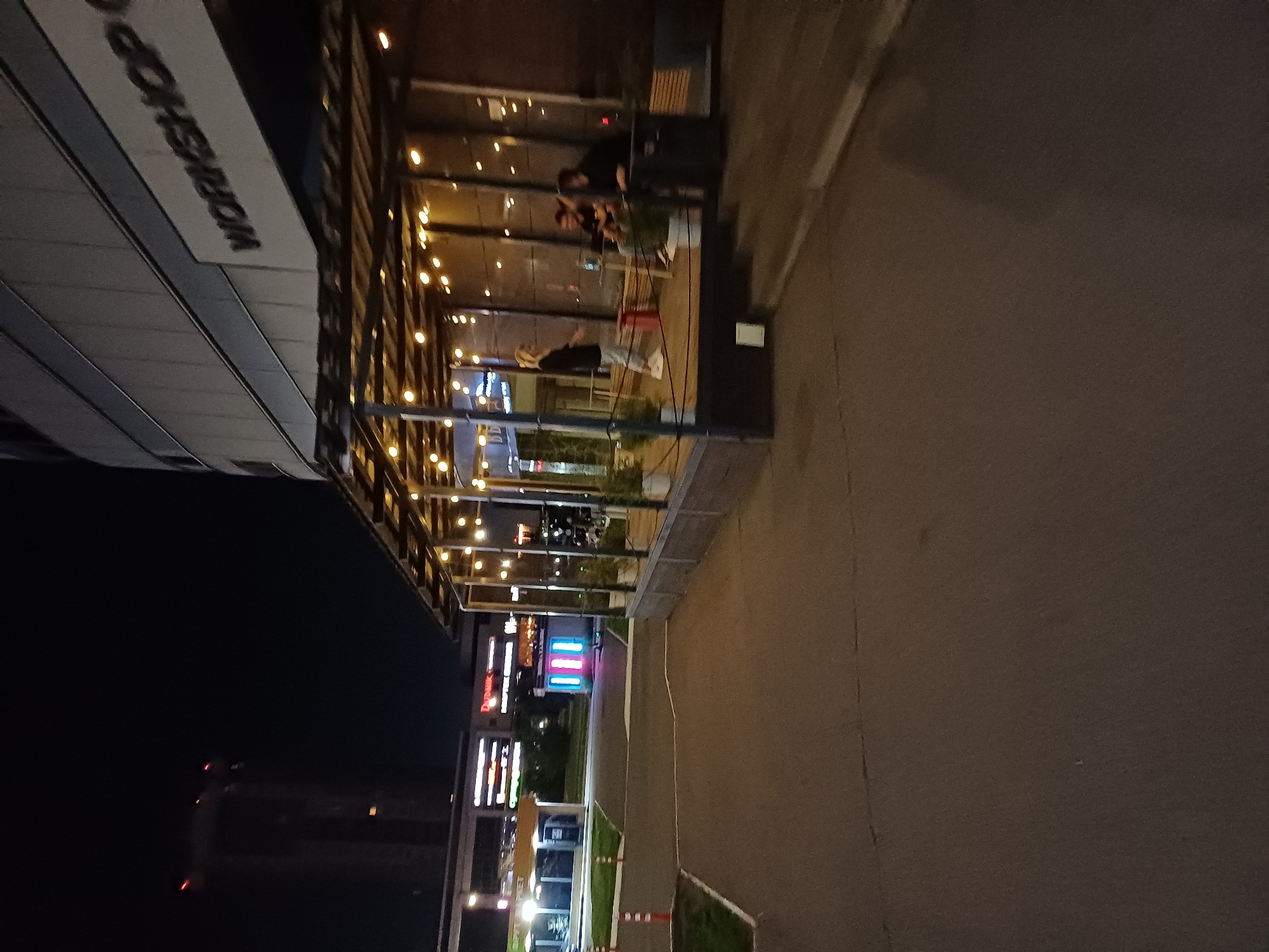 In a city setting at night, a multi-story building with a "WORKSHOP" sign is visible. In front of the building, an outdoor seating area with string lights is set up. Three people are seated at a table within this area. To the left, a street with a sidewalk and some buildings, including one with illuminated signs, can be seen. The overall scene is well-lit, with the lights from the building and seating area illuminating the surroundings.