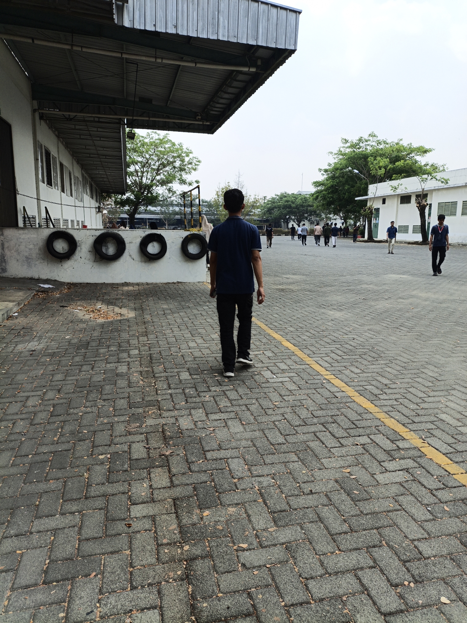 The image depicts an outdoor paved area, likely a parking lot or walkway, in front of a large white building with a corrugated metal roof overhang. The paving stones are rectangular and laid in a herringbone pattern. A yellow line is painted on the ground, dividing the paving.
In the foreground, a young man wearing a dark blue t-shirt and black pants walks away from the camera down the paved area. He has dark hair and is wearing white-soled sneakers.
In the middle ground, several people are walking in various directions. To the left, there is a low white wall with four black tires mounted horizontally. Beyond the wall, a portion of the white building is visible, with multiple windows. To the right of the building, another white structure with visible windows and some ventilation grates is present. Trees with green foliage are scattered throughout the scene.
In the background, more people are visible, and the sky is overcast and bright. The overall environment suggests an industrial or institutional setting. The image was taken in Nambangan, Indonesia.
