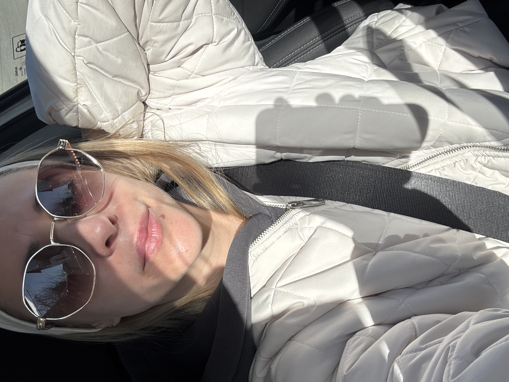 A woman wearing sunglasses and a cream-colored quilted jacket is sitting in what appears to be a car. She is wearing a seatbelt. The photo has strong light and shadow patterns. The location is not discernible as Chaikovsky, Russia from the image.