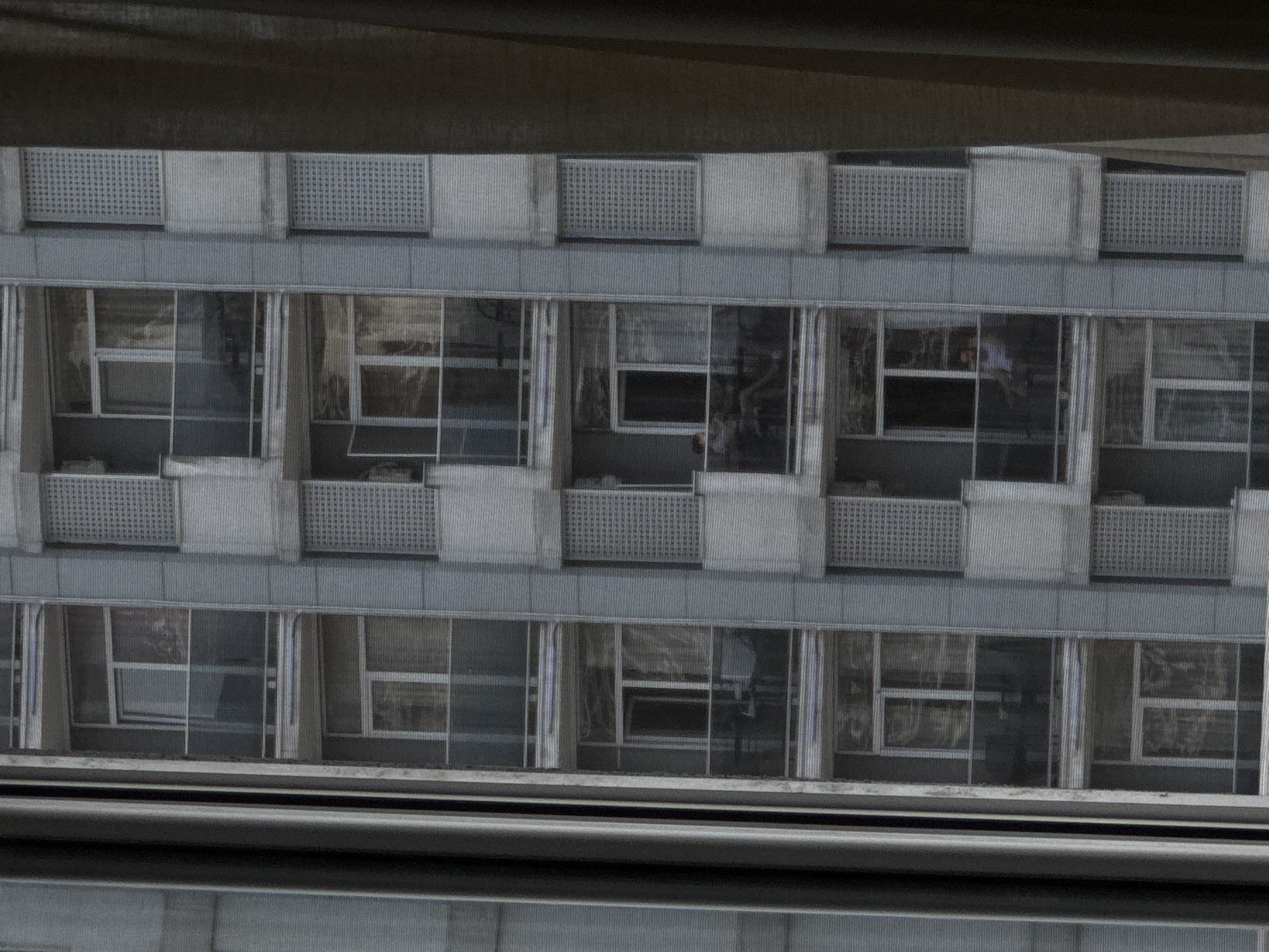 The media file shows a view of a building facade, likely a hotel or apartment complex, in Batumi, Georgia. The building features multiple stories with repeating patterns of windows and balconies. The windows are large and reflective, showing glimpses of interior spaces and possibly some furniture. Some balconies have decorative metal railings. The overall aesthetic suggests a modern, functional design. The lighting indicates it is daytime, but the sky and weather conditions are not clearly visible due to the angle and reflection. There is no visible text in the image. The scene is static, with no discernible activity or event taking place. The perspective suggests the image was taken from a higher vantage point, possibly another building or a drone, looking down at the facade. The colors are muted, dominated by grays and blues, consistent with a contemporary urban environment.