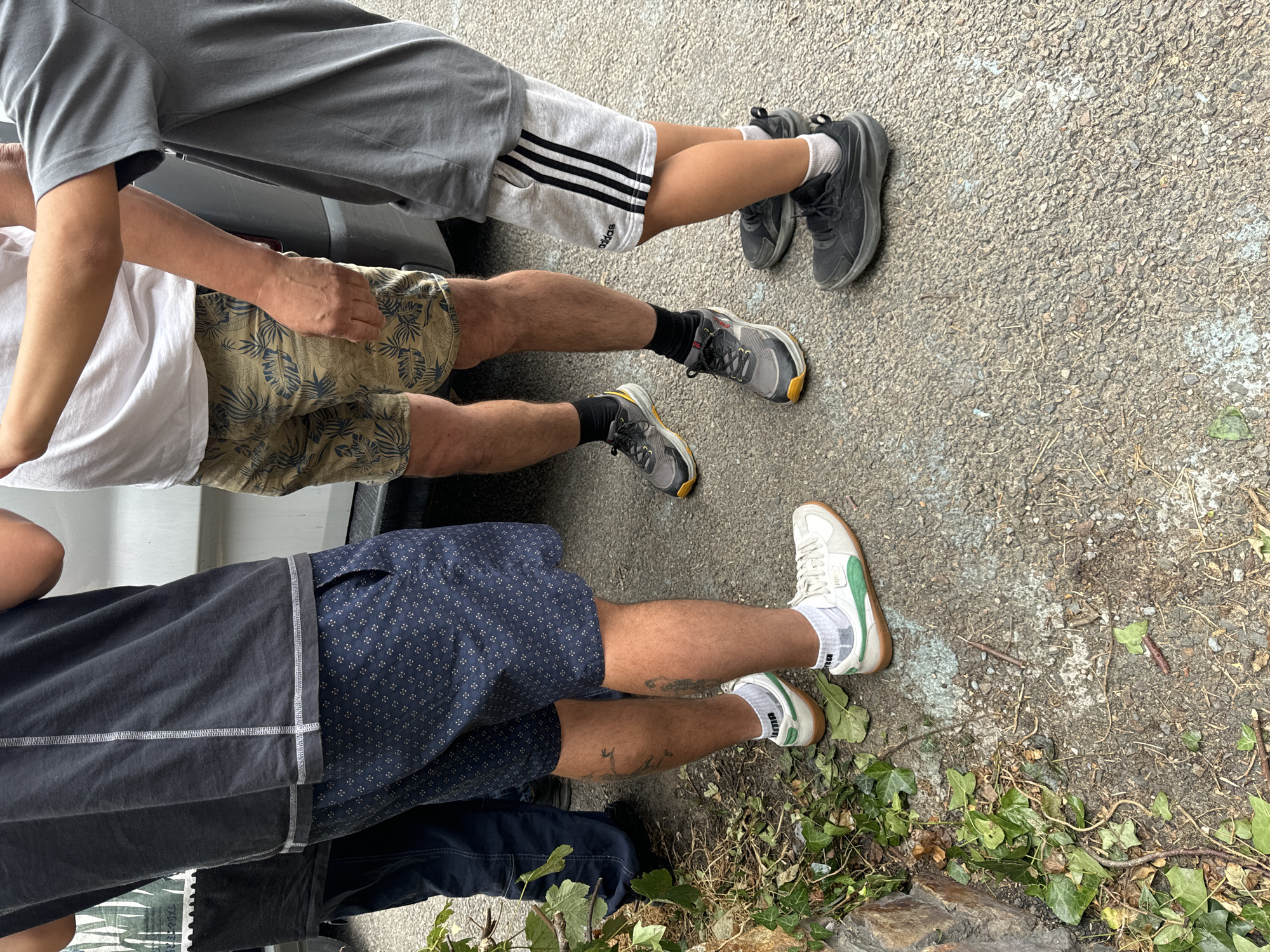 In Courtown, Ireland, four individuals stand on a paved surface. The lower halves of their bodies are visible, showcasing various attire. One person wears dark blue jeans and black shoes. Another is dressed in dark blue patterned shorts and white sneakers with green accents. A third individual is in light brown shorts with a leaf pattern and grey sneakers. The fourth person wears grey shorts with black stripes and the Adidas logo, along with black athletic shoes. One person has a tattoo of what appears to be a bird on their right calf. The ground is composed of asphalt with some patches of light blue paint and scattered debris including leaves and twigs.