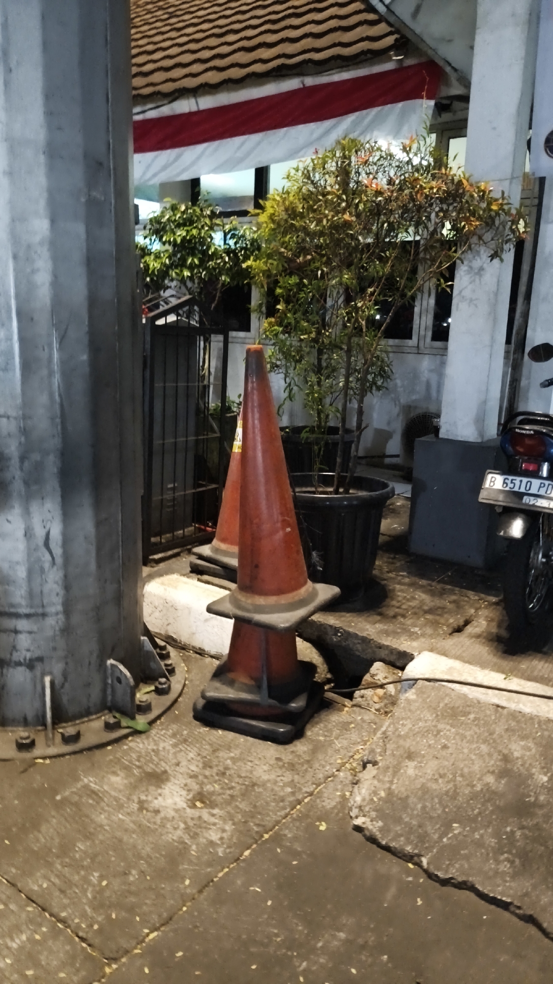 This image depicts a street scene at night, likely in South Jakarta, Indonesia, near a building with a tiled roof and a white and red banner. In the foreground, a large metal pole stands to the left, with two orange traffic cones placed in front of it. The cones appear to be stacked. Behind the cones, there is a black metal fence and several potted plants. To the right, a motorcycle is partially visible, with a license plate displaying "B 6510 PD." The ground is paved with concrete slabs, some of which appear cracked or uneven. The lighting suggests it is nighttime, with artificial lights illuminating the scene. There are no people visible in the image.