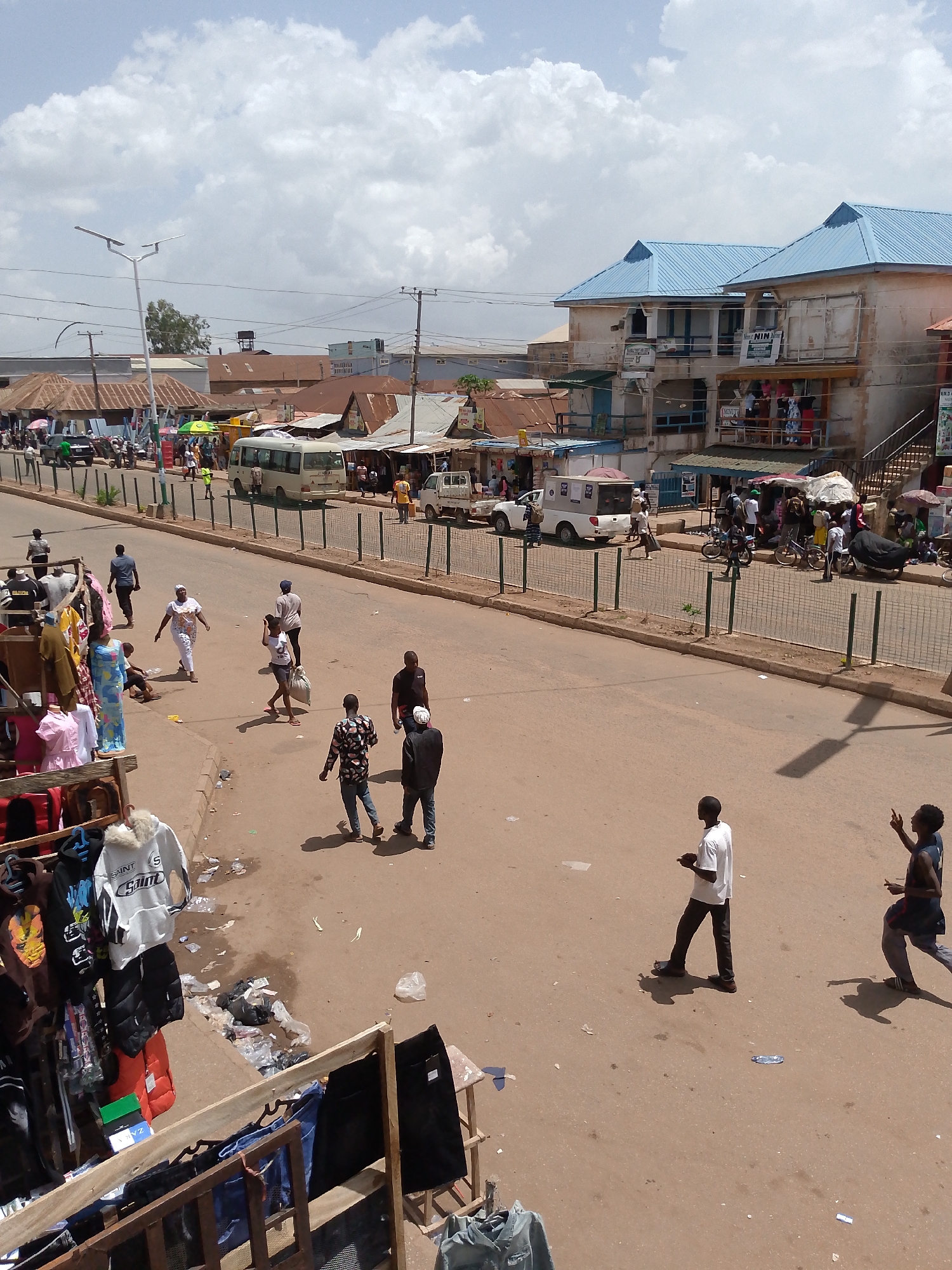 The image depicts an elevated view of a street scene in what appears to be Bukuru, Nigeria. A street runs horizontally across the frame, separating buildings and businesses on one side from a commercial area with clothing racks on the other.
Several pedestrians are walking across the street, including two individuals in the lower right corner, one wearing a white shirt and the other a blue sleeveless shirt. Two men in patterned shirts and dark pants cross the street further up. A woman in white shirt and skirt can be seen in the middle of the street with a shopping bag.
Various vehicles, including a white van, white pickup, and taxis, are parked along the roadside. Makeshift stalls and umbrellas indicate a marketplace activity on the far side of the street. The buildings across the street are two stories high, with one building exhibiting a distinctive blue roof. A metal fence runs along the edge of the sidewalk. There is visible litter and debris on the street surface. The sky is partly cloudy.