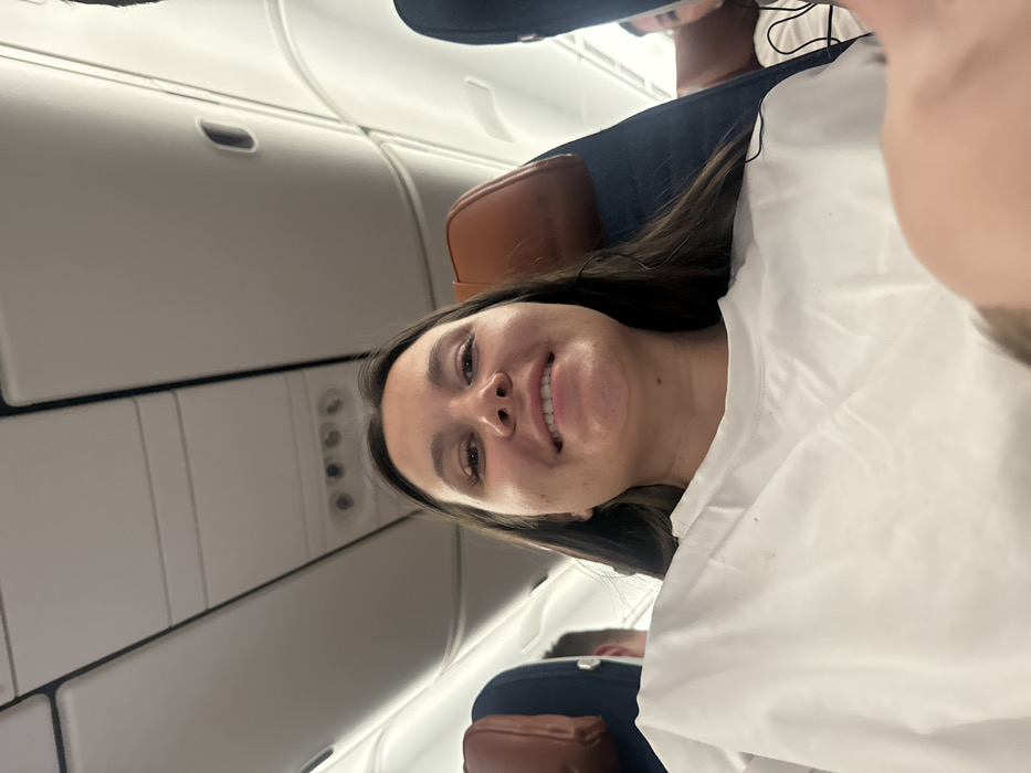 The image depicts a young woman posing for a selfie or being photographed inside an airplane cabin. She is positioned prominently in the foreground, looking directly at the camera with a wide, joyful smile. Her long, dark brown hair is visible around her face, and her dark eyes are bright. She is wearing a simple white top, and a white blanket or sheet is draped over her, covering her chest and left shoulder. Her head appears to be resting against a seat or pillow.
The background reveals the interior of an airplane. Above and to the left of the woman's head, white overhead compartments are visible, along with a panel featuring air vents and passenger service lights. Below and to the right of the woman, another passenger is partially visible, appearing to be a male. This individual is wearing a dark blue garment, possibly a shirt or jacket, and is slumped back in their seat, seemingly resting or sleeping. A brown headrest, possibly leather or faux leather, is visible behind both the main subject and the other passenger. A thin, dark wire, possibly from headphones, can be seen near the second passenger's shoulder.
The lighting within the cabin is artificial, contributing to a slightly dim but well-lit appearance around the main subject. The overall atmosphere conveyed is one of relaxation and contentment, largely due to the woman's cheerful expression. No discernible text is visible in the image.