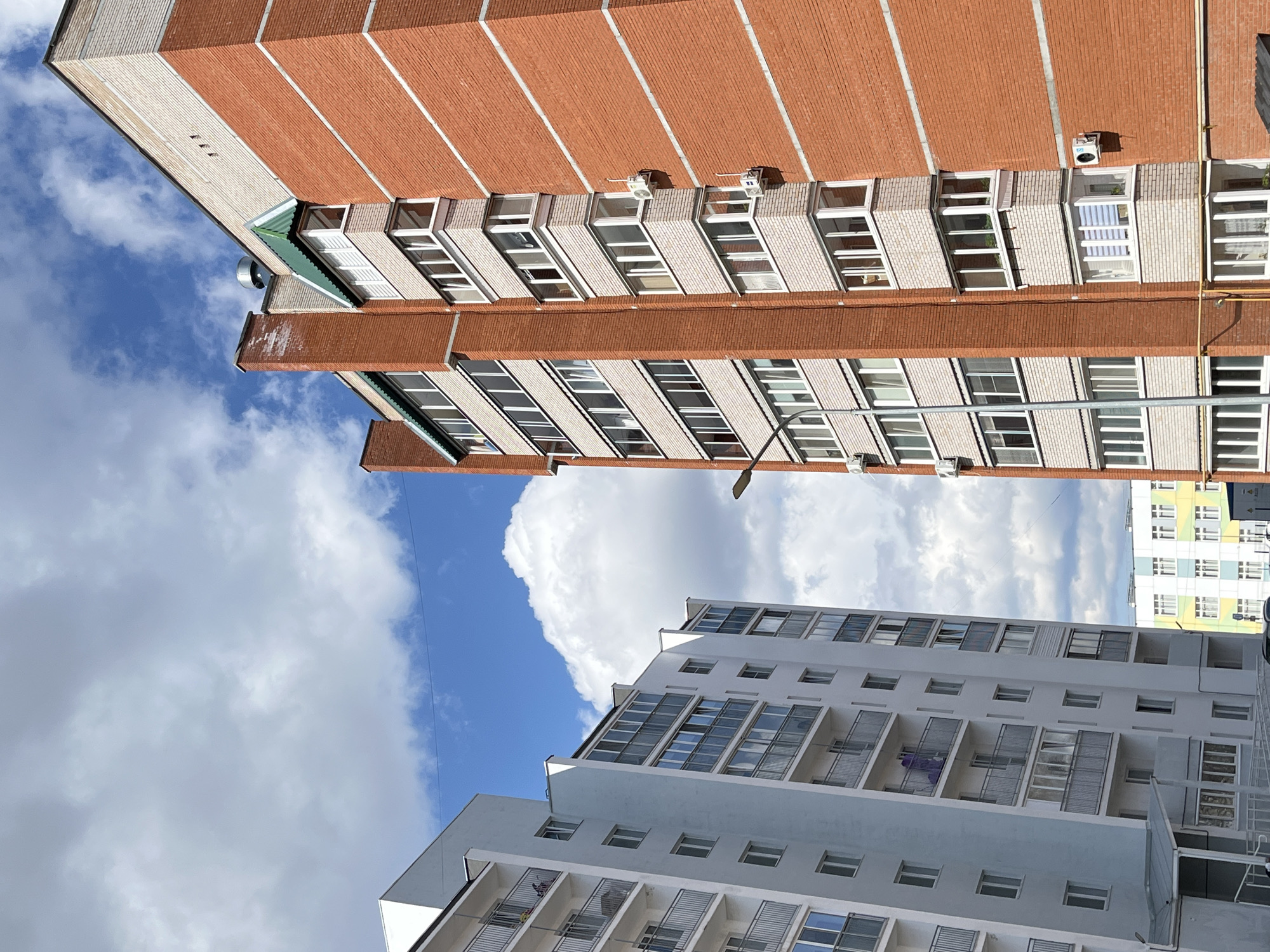 The image presents a rotated view of an urban landscape in Chaikovsky, Russia, featuring multiple residential buildings against a bright, cloudy sky. The photograph is taken from a low angle, looking upwards, and is rotated 90 degrees clockwise, giving the impression that the structures are leaning.
The most prominent building, occupying the upper and left parts of the frame, is a multi-story residential complex constructed with alternating bands of red brick and lighter-colored (possibly cream or white) masonry. It features numerous windows, some with enclosed balconies or sunrooms, and several external air conditioning units. A distinctive green-colored roof or awning is visible near the top-left corner, and a yellow gas pipe runs vertically along one side of the building.
Below and to the right, a second, more modern residential building in shades of white and grey rises. This building showcases balconies with metal railings, and on one balcony, purple laundry is visibly hanging to dry. Its design is characterized by multiple rows of windows and a tiered structure. In the far background, to the right of the white building, a third residential building with a colorful, checkered facade of yellow, green, and white panels is partially visible.
The sky above is a vibrant blue, dotted with large, fluffy white cumulus clouds, indicating a clear and sunny daytime. A streetlamp fixture is discernible in the mid-ground, extending upwards between the buildings. No visible text or discernible signage appears on any of the buildings or objects. The scene captures the varied architecture of a modern Russian city against an open sky.