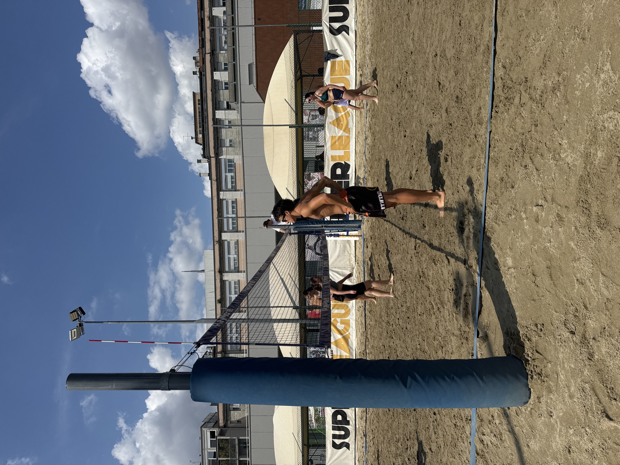 This image depicts an outdoor beach volleyball court located in Milan, Italy, under a clear blue sky with scattered white cumulus clouds. The court surface is light brown sand, showing impressions from movement.
In the immediate foreground, a padded, dark blue cylindrical volleyball net post stands vertically on the left, partially obscuring the court. A dark blue boundary line (tape) is visible on the sand, extending from the base of this post to the right.
Four individuals are visible on the court, all barefoot.
1. A shirtless male, with fair skin and dark hair, stands prominently in the mid-left foreground. He wears dark shorts with a white "MMA" logo on the left leg and dark sunglasses. He is facing left with his hands on his hips, head tilted slightly upwards. His shadow is cast towards the right.
2. Behind the net on the left, two females are present. One female, with dark hair, wearing a dark athletic top and shorts, stands facing away from the camera, positioned near the net. Another female is partially visible behind her.
3. On the right side of the net, another female, with dark hair, wearing a green sports bra and dark shorts, walks towards the right edge of the frame.
The volleyball net itself is dark and stretches across the court. A white and red striped antenna is attached vertically to the net on the left side, extending upwards. A tall, grey pole with two floodlight fixtures is positioned behind the net on the left.
In the midground, beyond the court, a white banner with dark text and yellow sections, reading "SUPER LEAGUE" repeatedly, spans the width of the scene. Behind the banner, various urban structures are visible:
* On the left and center, multi-story buildings with numerous windows, some featuring balconies or industrial elements, are present.
* Towards the right, a large, light-colored tensile structure or canopy tent is erected, providing shade.
* Fencing, likely green, separates the court area from the background structures.
* A fifth person, wearing a white shirt, is partially visible standing near a net post in the far background on the left, beyond the main court area.
The scene is brightly lit by direct sunlight, evidenced by the sharp shadows cast on the sand and objects.