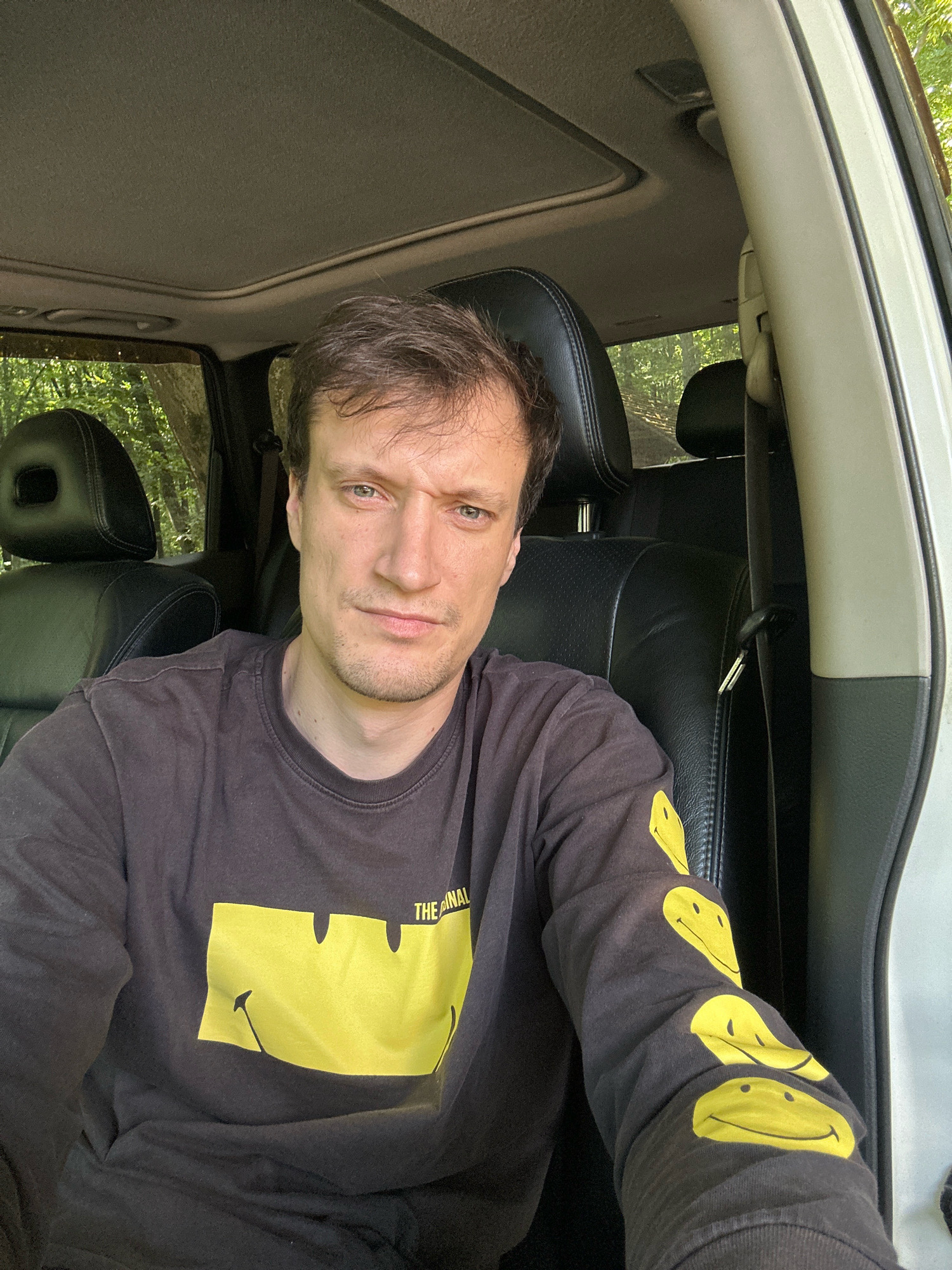 A man with light skin, brown hair, and a blue-eyed gaze is seated inside a vehicle. He is wearing a black long-sleeved shirt with a yellow smiley face graphic and the word "THE FINAL" printed on it. The man's expression is a neutral grimace. The interior of the car features black leather seats and a sunroof. Outside the car, green trees are visible, suggesting a natural, possibly rural or wooded setting. The image does not contain any information about the city of Shatava, Ukraine.