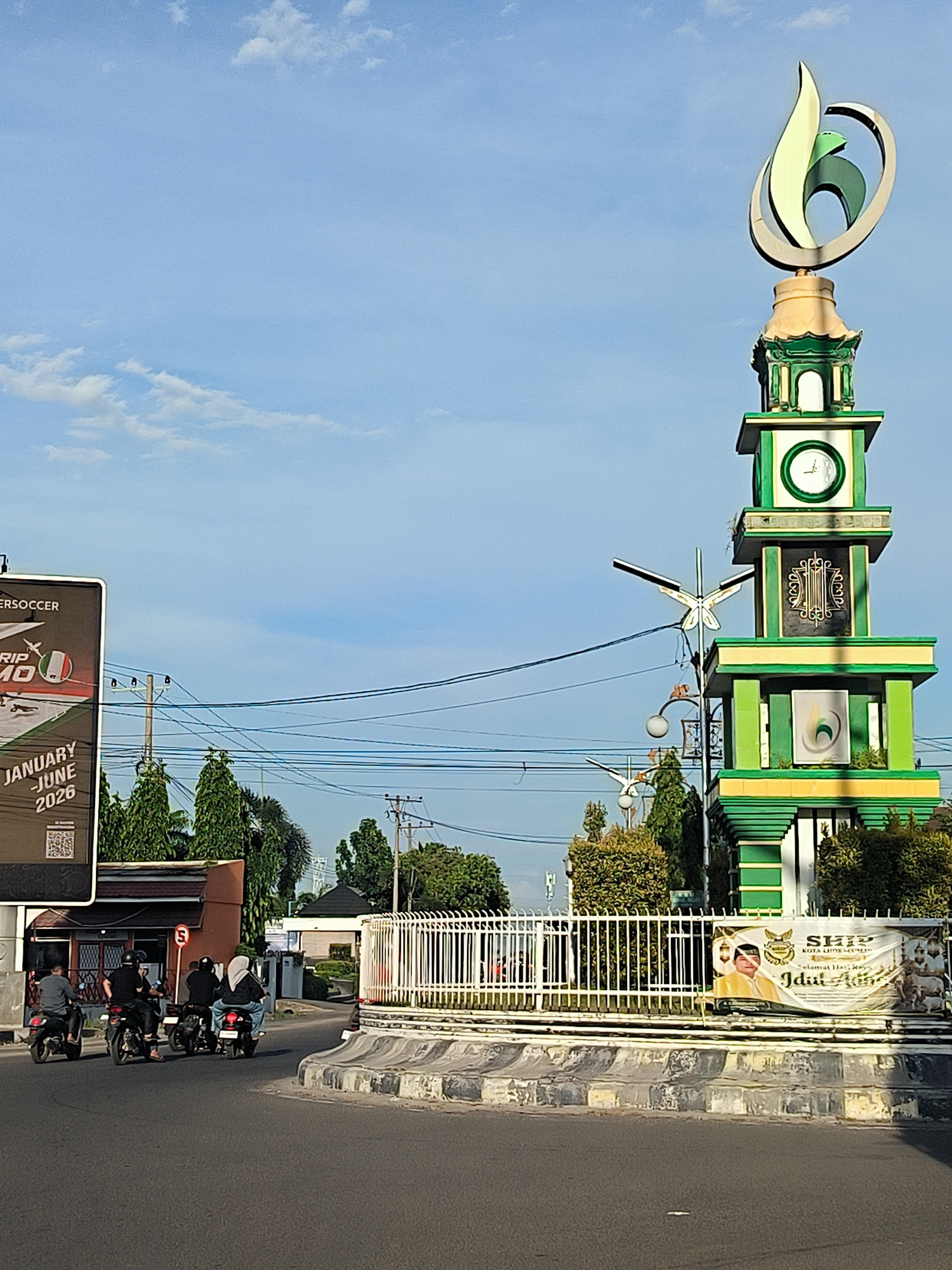 A street scene in Lhokseumawe, Indonesia, features a roundabout with a monument resembling a clock tower topped with a stylized green and silver logo. The tower is constructed with tiers of green and white blocks, featuring a clock face, decorative grillwork, and greenery. A white fence surrounds the raised roundabout base, which is adorned with a banner featuring an individual in a yellow garment, with text in Indonesian, suggesting a religious or cultural celebration. Four motorbikes, each with a rider, are driving on the dark asphalt road toward the right side of the frame. A large billboard advertising "BERSOCCER TRIP MO" and the dates "JANUARY-JUNE 2026" is seen to the left. Overhead are power lines spanning across the scene. The sky is bright blue with scattered clouds. Buildings and trees are visible in the background.
