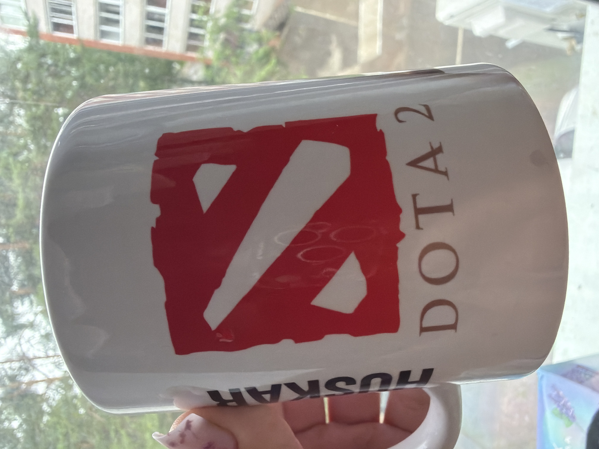 A close-up shot captures a white mug with the Dota 2 logo emblazoned on it. The logo is a stylized red "S" shape. Vertically oriented text reading "HUSKAR" is printed on the left side of the mug, with the letters in black. The text "DOTA 2" is printed horizontally below the logo in a brown font. A hand with painted fingernails is visible holding the mug, with the thumb and index finger gripping the handle. The mug is positioned in front of a window with a view of trees and a multi-story apartment building, suggesting an urban setting. Based on the architecture, the location is likely Chaikovsky, Russia.