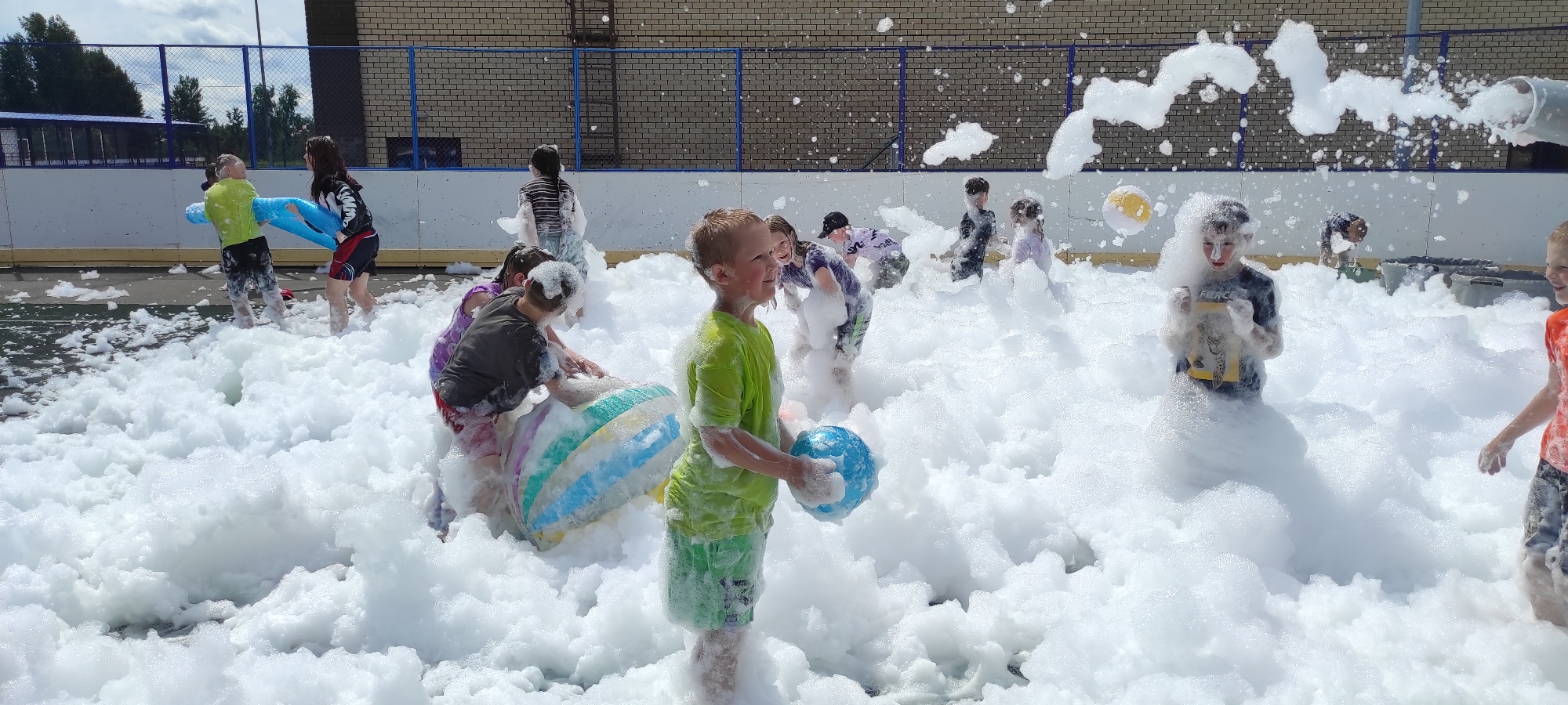 This image depicts a group of children engaged in a foam party in Noviy, Russia. The scene is dominated by a large quantity of white foam, suggesting a recreational event likely held outdoors, possibly at a park or schoolyard, given the presence of a brick building and a fence in the background.
Several children are visible, mostly partially submerged in the foam. Their clothing is varied, with some wearing bright colors and others darker tones. Some children are holding or interacting with inflatable toys, including a large, multi-colored inflatable ball and a blue inflatable object that resembles a dolphin or a tube. One child in the foreground, wearing a green t-shirt and shorts, is holding a blue ball. Another child in a dark shirt and black shorts is covered in foam, with a playful expression. To the left, two children are seen struggling with a blue inflatable toy. In the background, more children are discernible amidst the foam, some appearing to be running or playing.
The environment appears to be an open area with a paved surface visible beneath the foam in some sections. The sky is partly cloudy, indicating daytime. The overall atmosphere conveyed is one of fun and energetic play, with the foam creating a playful and slightly chaotic environment for the children's activities.