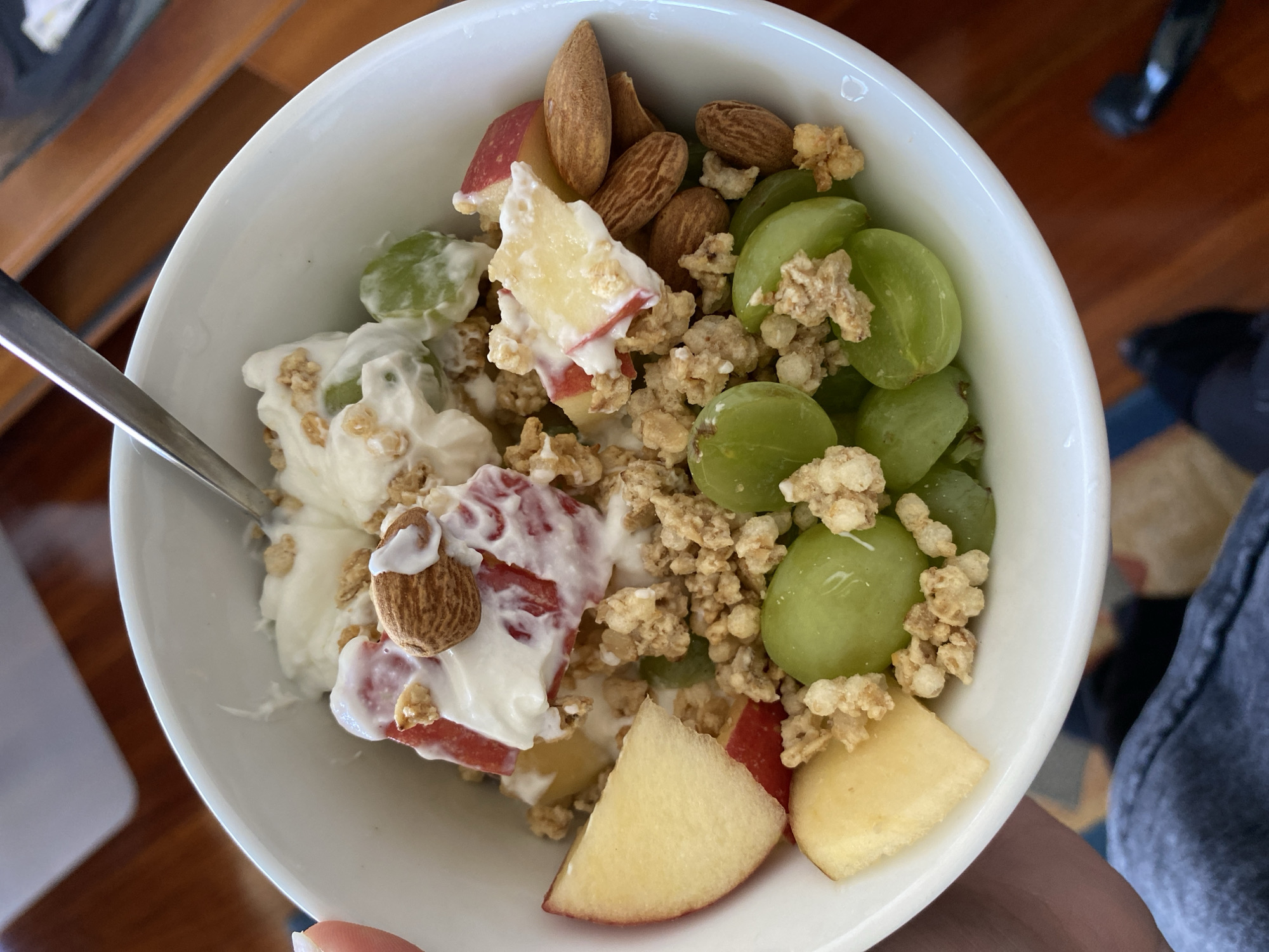 A top-down, close-up shot shows a white bowl filled with food. The bowl contains green grapes, sliced apples (some with red skin, some without), yogurt, granola, and almonds. A silver spoon is partially submerged in the yogurt on the left side of the bowl. The bowl is being held by a hand with visible fingernails. The background shows a wooden surface, possibly a table, and a glimpse of a blue and patterned rug. The scene appears to be indoors. The location in Gijón/Xixón, Spain cannot be determined from the image.