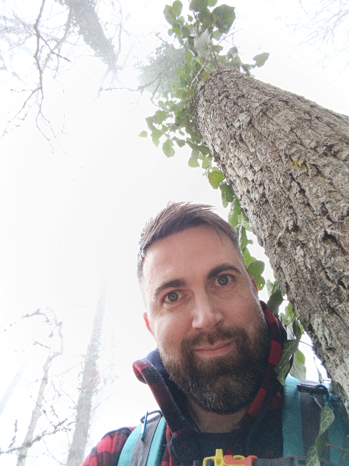 An upward-angled portrait depicts an adult male in a foggy forest environment located in Гурзуфский поселковый совет, Ukraine. The subject, positioned in the lower and left-center of the frame, has short brown hair, a full dark beard, and blue-green eyes, looking directly at the camera with a slight smile. He is wearing a red and black plaid hooded garment and a turquoise backpack with a yellow and orange buckle visible on the front strap. A large tree trunk with textured bark and green leaves growing on it ascends vertically along the right side of the image. The background is characterized by dense white fog or mist, through which the silhouettes of additional bare tree branches are faintly discernible.