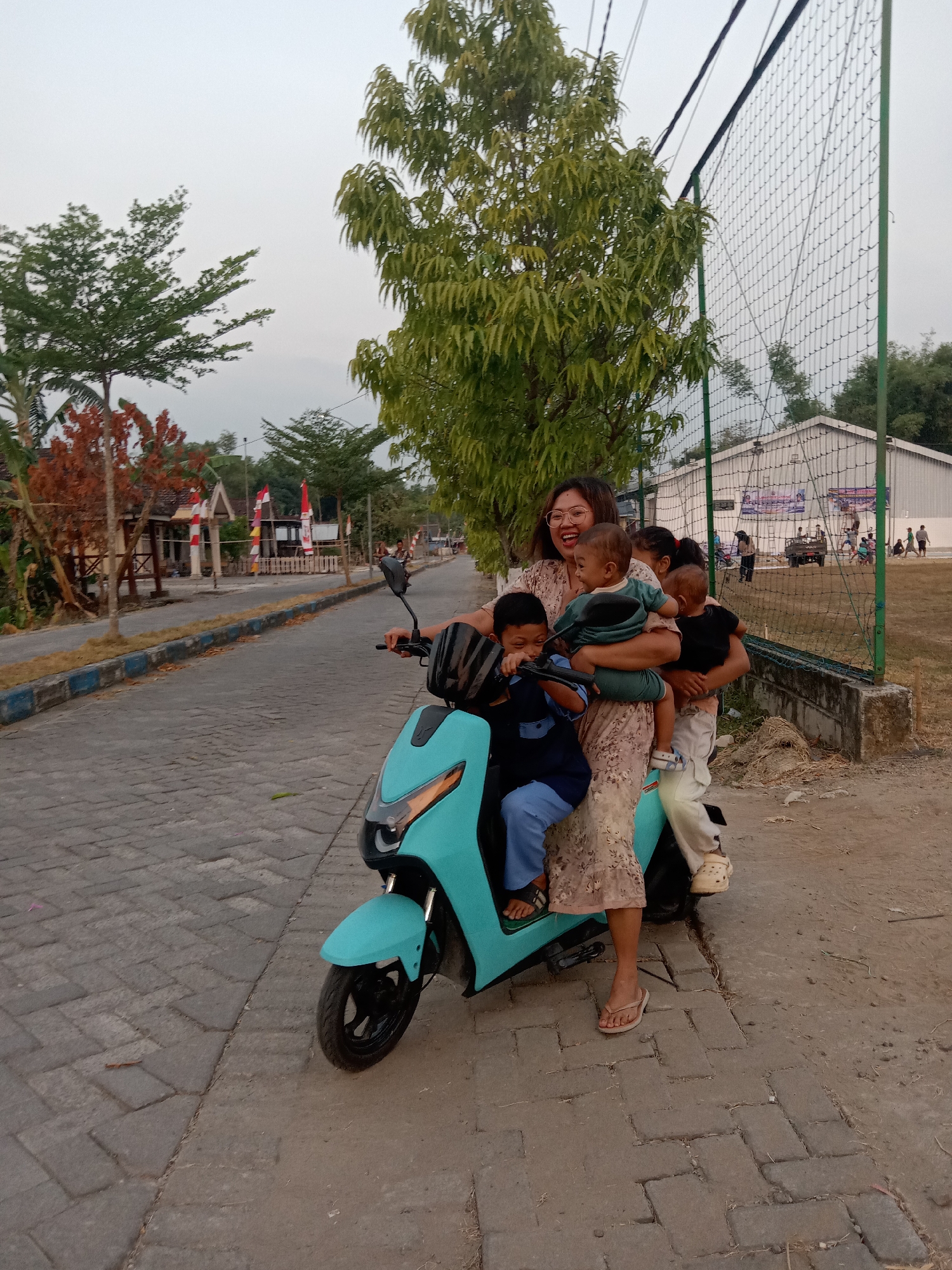 A woman and four children are on a light blue electric scooter on a paved street in Stiaji, Indonesia. The woman, wearing a floral dress and glasses, is in the middle, holding two babies. One child is in front of her, and another is seated behind her. The scooter has a black helmet on the handlebars. The street is lined with trees and buildings. In the background, there's a sports field with a fence and people playing. The sky is overcast.