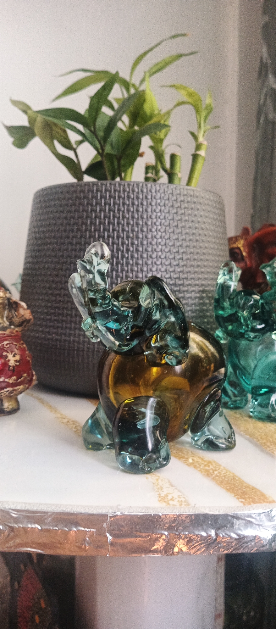 A close-up, eye-level, indoor shot focuses on a translucent, amber and teal glass elephant figurine. The elephant is positioned centrally, facing left, with its trunk raised and its ears spread wide. Its body is a smooth, rounded amber sphere, and its legs and trunk are made of intricately molded teal glass. The figurine rests on a white surface adorned with a shimmering, gold striped pattern. Behind the elephant, a dark gray, woven planter holds a vibrant green bamboo plant. To the right of the elephant, another teal glass figurine, possibly another elephant, is partially visible. To the left, a small, red and gold decorative object is also in the frame. The background is a blurred white wall, suggesting the items are displayed on a shelf or table.