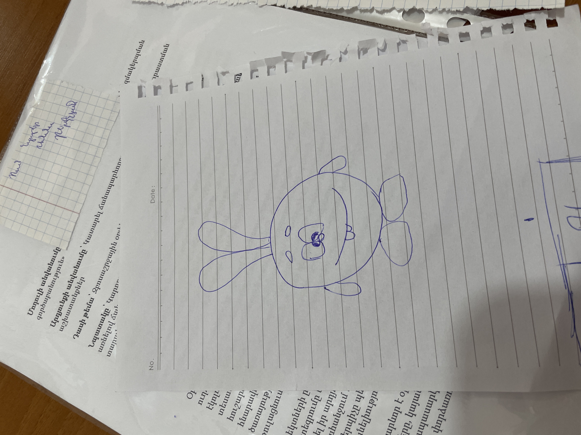 The image shows a lined piece of paper with a blue ink drawing of a cartoon-like character with large eyes and ears, presumably a child's drawing. The paper also has some Armenian text printed on it. Adjacent to this paper are other documents, also in Armenian. The setting appears to be indoors, possibly on a desk or table. There's no indication of the location in Yerevan, Armenia, or any people, actions, or interactions happening.