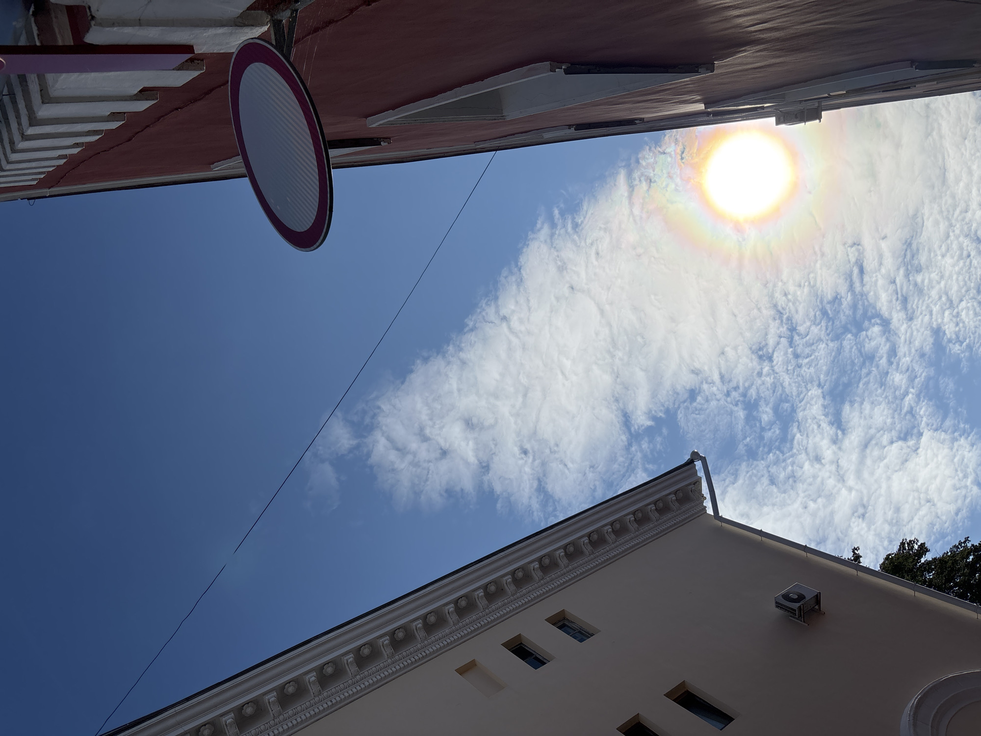 The image captures a low-angle view of a street in Homyel, Belarus, with two buildings flanking a bright blue sky filled with wispy clouds. The building on the left appears to be a light-colored structure with ornate trim along its roofline and several rectangular windows. An air conditioning unit is mounted on its exterior wall. The building on the right is a taller, reddish-brown structure with a decorative white cornice and what appears to be a cracked facade. A circular traffic sign with a pink border and a white, striped interior is mounted on the reddish building. The sun is prominently visible in the sky, creating a halo effect with a rainbow-like luminescence, suggesting it's either midday or late afternoon. A thin wire stretches across the sky, connecting to unseen points beyond the frame. A dark green tree is partially visible at the bottom left of the image.