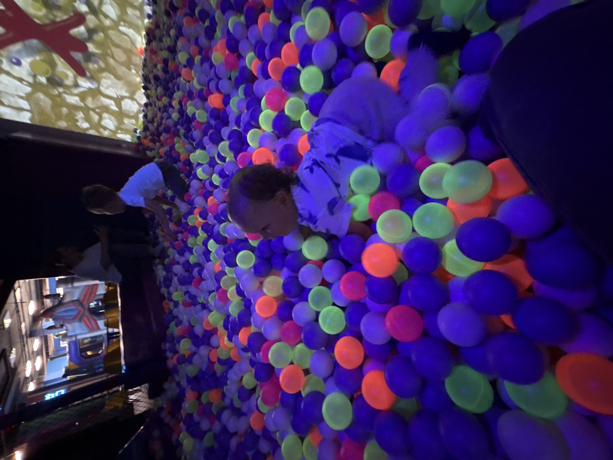 The image shows a room filled with colorful balls, mainly purple, orange, and yellow. A young child in a light-colored shirt is partially submerged in the ball pit. In the background, a screen displays an image, possibly of a city scene with a building resembling a toy version of a construction. It appears to be a play area, with the scene suggesting that it could be located at a children's entertainment center in Izhevsk, Russia. People can be seen in the background appearing to be playing in a ball pit.