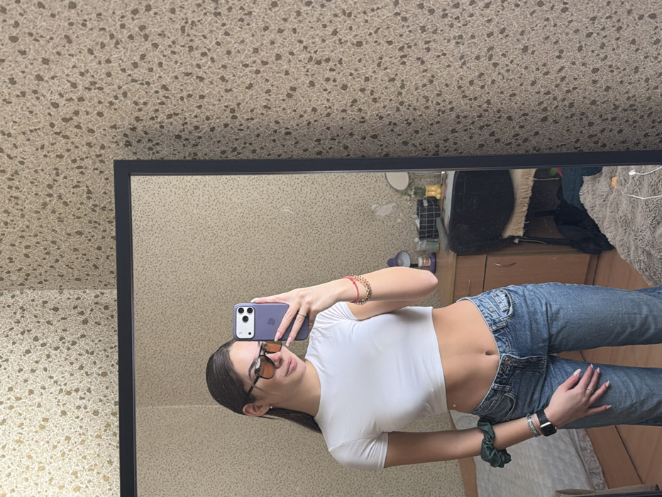 The image displays a mirror selfie of a female individual taken inside a private residence in Sofia, Bulgaria. The subject, positioned centrally in a black-framed rectangular mirror, is wearing a white short-sleeved cropped top, light-wash low-rise jeans, and amber-tinted rectangular sunglasses. Her dark hair is pulled back. The subject's left hand, adorned with a ring on the ring finger, holds a purple smartphone with a light grey camera module, used to capture the image. A green scrunchie, a black smartwatch, and multiple bracelets are on her left wrist; red and gold chain bracelets are on her right wrist. The reflected environment reveals a bed to the left, and a brown wooden nightstand to the right. On the nightstand, a purple desk lamp, a small white bottle, a black wire basket, and a small round mirror are visible. The floor is partially covered by a light-colored, textured rug, on which a white charging cable rests. The room's walls are covered in a light beige, speckled wallpaper.