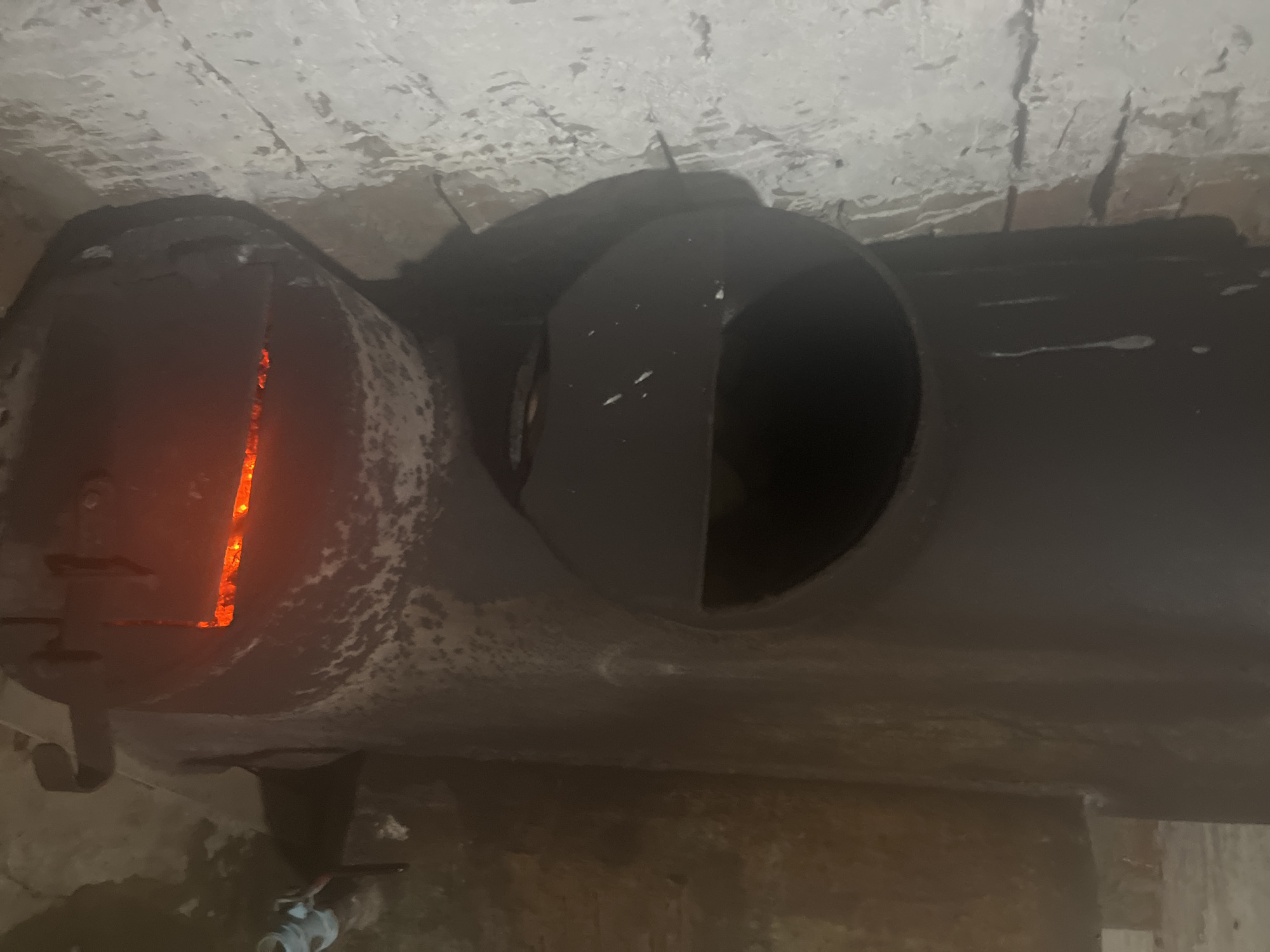 The image shows a wood-burning stove with glowing embers visible through an opening at the bottom. The stove is dark-colored and appears to be made of metal. It is positioned in a room with rough, light-colored brick walls. To the right of the stove, there is a glimpse of a brown, textured surface, possibly another wall or object. The overall scene suggests an indoor environment, likely a utilitarian space. The location is specified as Уральское, Russia.