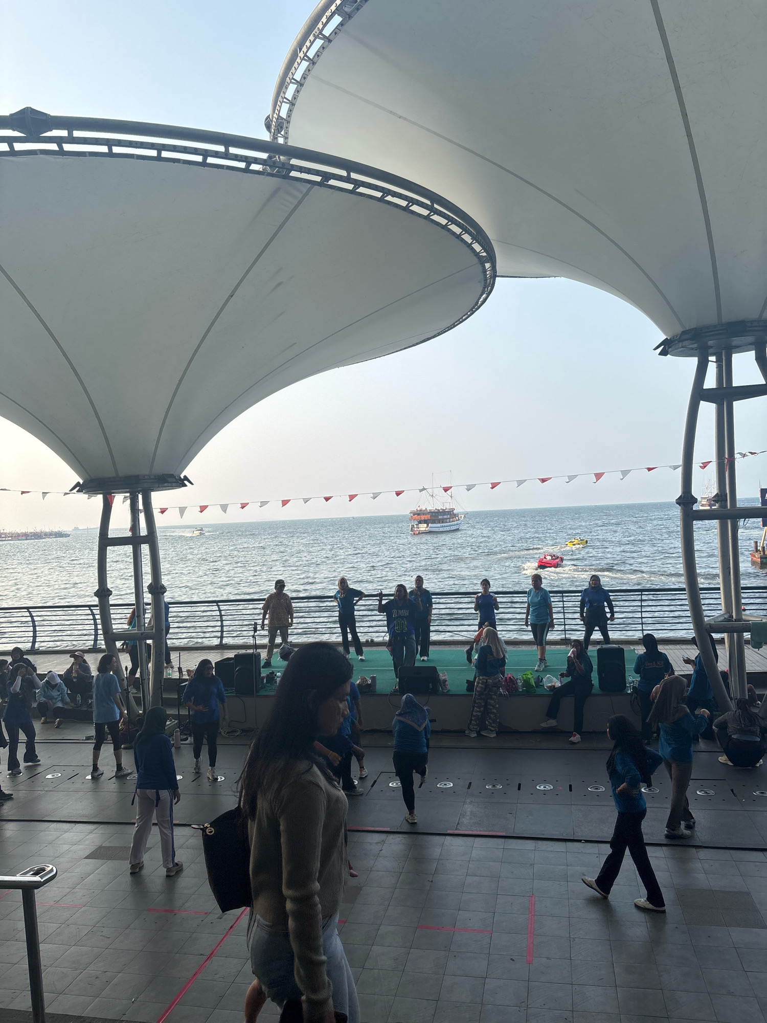 The image depicts an outdoor event near the Ancol beachfront in North Jakarta, Indonesia. Underneath a large, white, tensile fabric canopy, a group of people are engaged in what appears to be a dance or fitness session on a raised stage. Several individuals are actively participating in the activity, while others are seated or standing around the periphery, observing. The background reveals a calm sea with a few boats, including a larger sailing vessel and two smaller speedboats creating wakes. Decorative flags are strung across the railing separating the waterfront promenade from the sea. The overall atmosphere suggests a public gathering or recreational event.