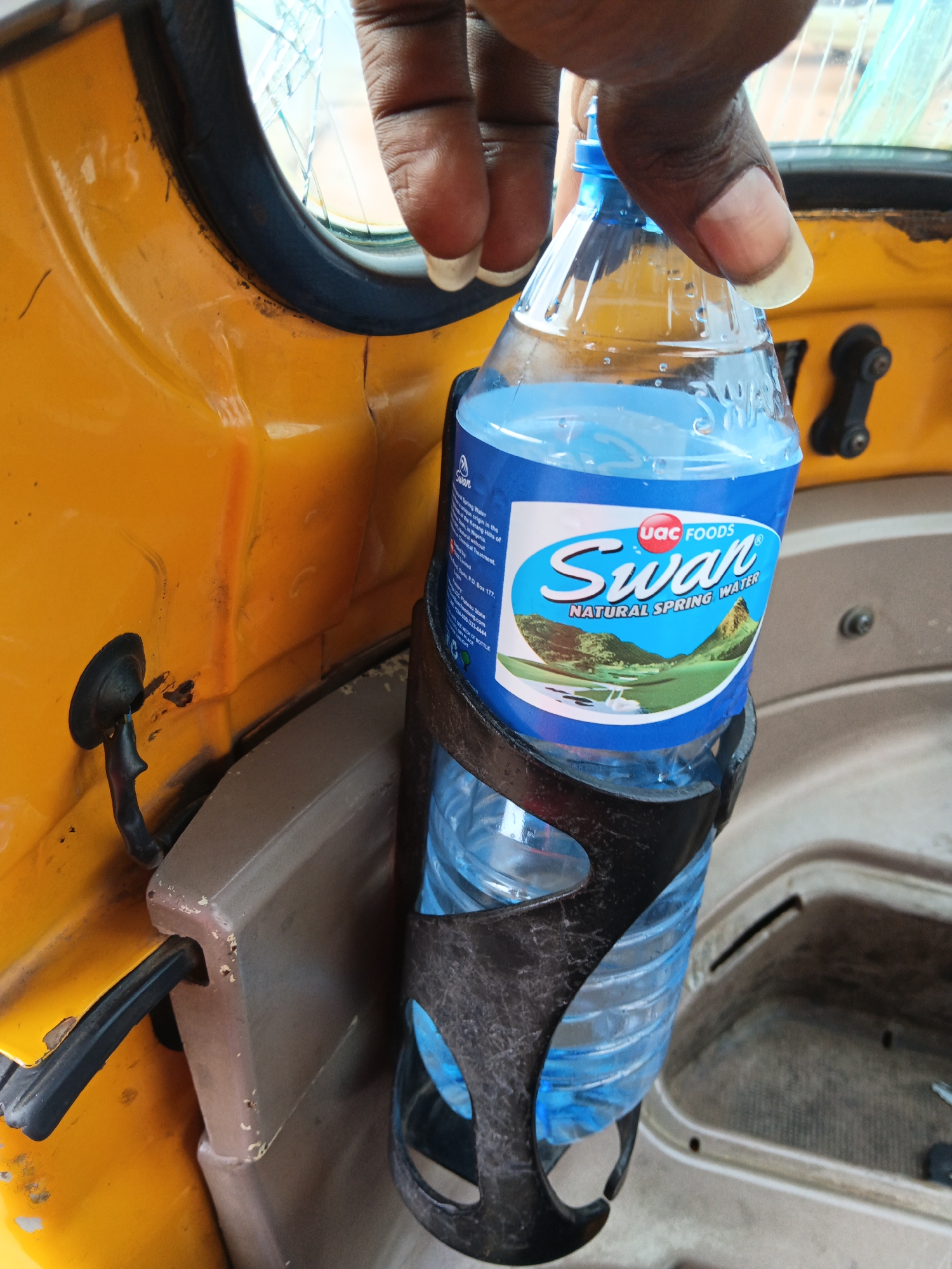 A person's hand holds a plastic bottle of Swan Natural Spring Water, positioned in a black plastic cup holder inside a yellow vehicle. The vehicle interior appears to be part of a tricycle or auto-rickshaw, with visible yellow painted bodywork and a dark grey console or dashboard. The bottle has a blue label with the Swan logo, depicting a pair of swans on a lake with mountains in the background. The vehicle's windshield is cracked. The scene is captured from a medium close-up perspective, focusing on the bottle and the hand. The background is blurred, indicating a shallow depth of field.