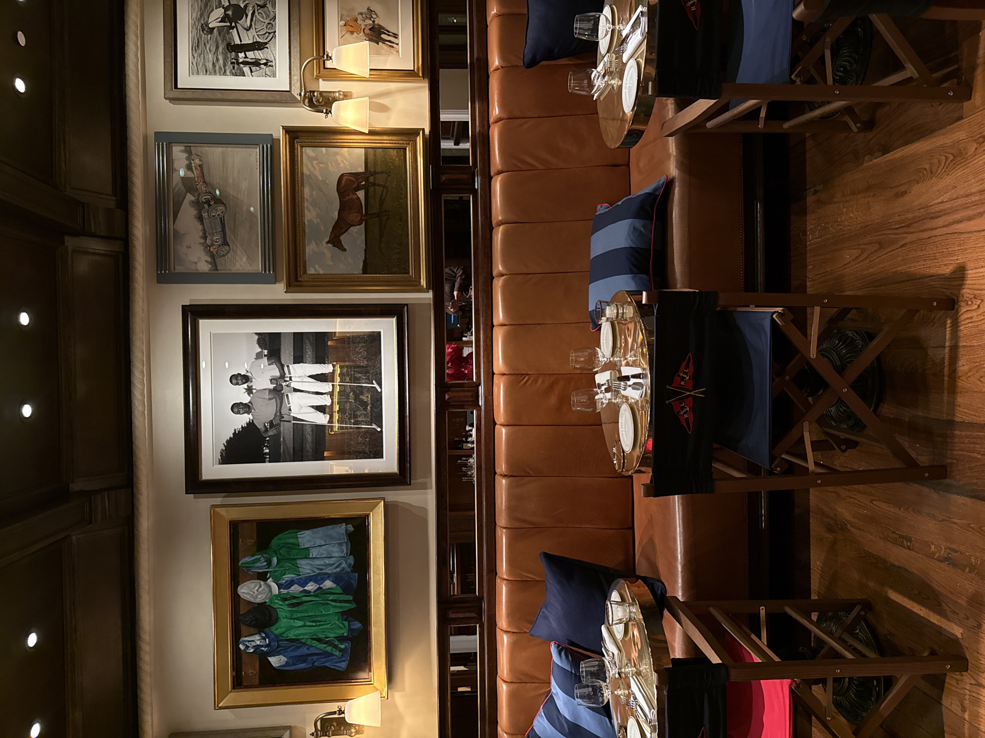 The image depicts a restaurant interior. The scene consists of rows of wooden chairs placed around small, round tables set for dining, complete with silverware and glassware. The chairs are upholstered in blue fabric, and some have decorative pillows. The tables are placed against a long, brown leather banquette.
On the wall opposite the banquette, there is a collection of framed artwork. The paintings and photographs include equestrian themes, racing scenes, and portraits. The walls are painted in a light neutral color.
The restaurant appears to be well-lit and has a formal yet cozy ambiance. Based on the style of the interior decoration and the presence of Ralph Lauren branded items on the seat backs, it can be inferred that this is the restaurant Ralph's Milan, in Milan, Italy.