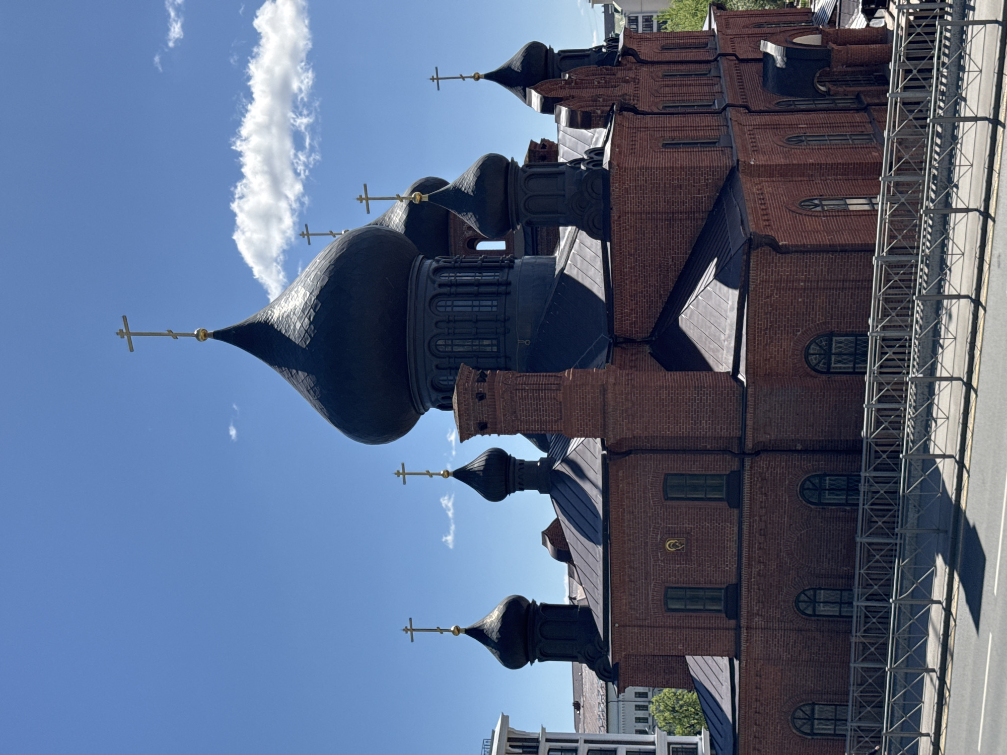 The image shows a brick building with several dark domes topped with golden crosses. The architectural style suggests an Eastern Orthodox church. A long, slender white cloud is visible in the blue sky behind the domes. Metal scaffolding runs along the side of the building adjacent to what appears to be a street or road. The location is Kazan, Russia, based on the architectural characteristics of the building and its domes.