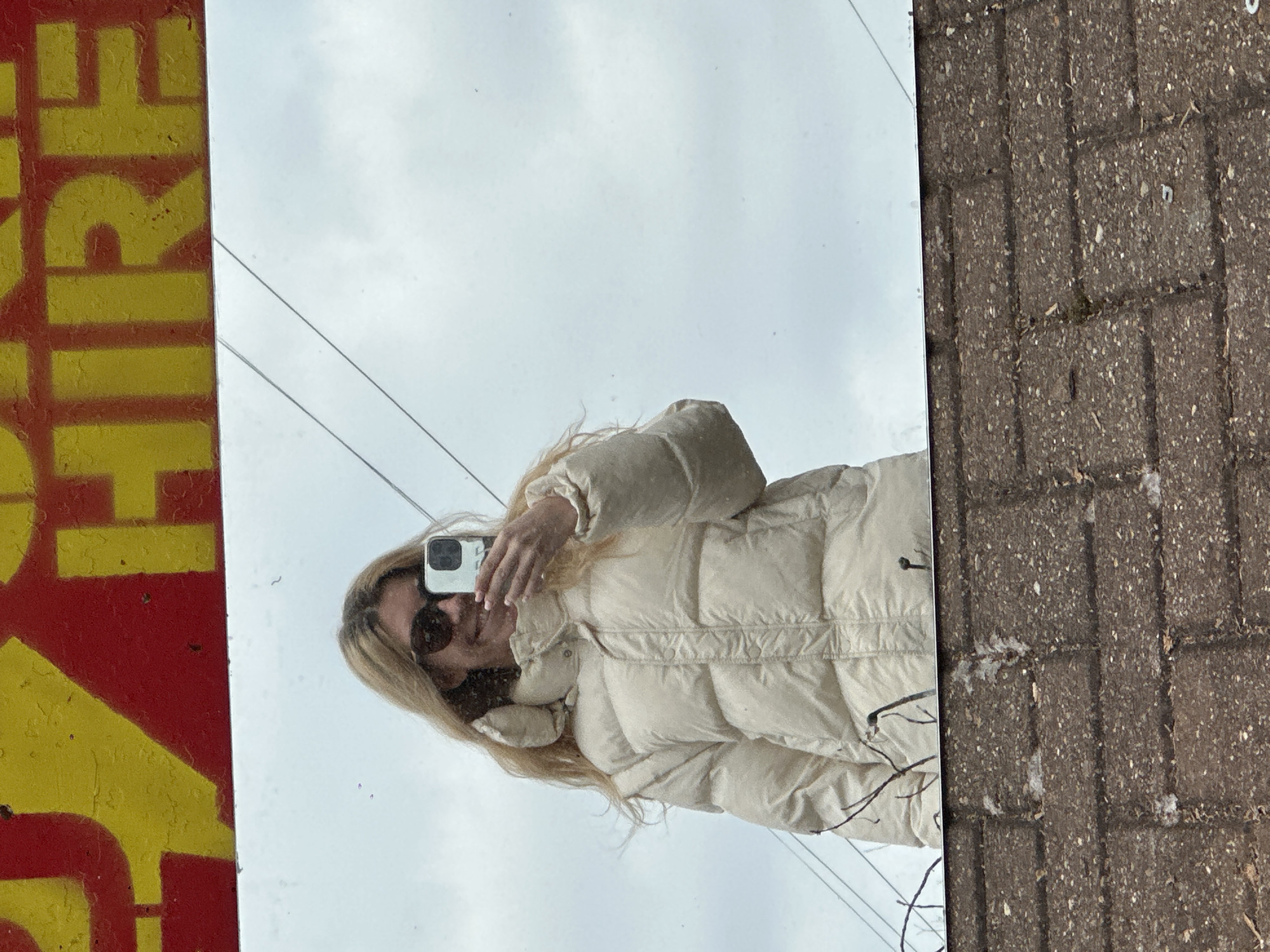 A person wearing a beige puffer jacket and sunglasses is reflected in a mirror held up against a red and yellow wall on the left and a brick wall on the right. The reflection shows the person holding a smartphone up as if taking a selfie, with a cloudy sky in the background. The location is London, United Kingdom.