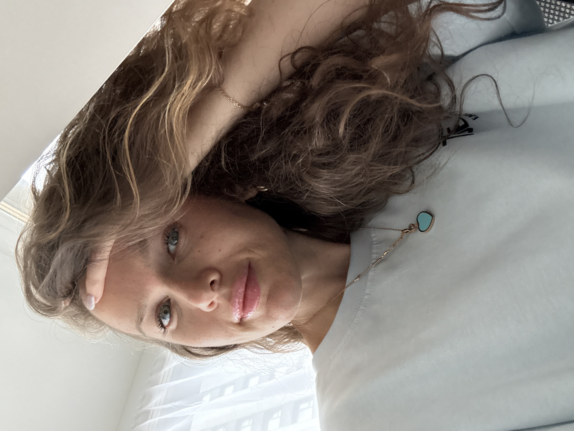 The image is a close-up selfie of a young woman with long, wavy brown hair. She is wearing a light blue t-shirt and a necklace with a teal heart-shaped pendant. Her left hand is resting on her forehead, and her right arm is behind her head. The background appears to be an indoor setting with white sheer curtains. There is no information in the image to suggest the location is Moscow, Russia.