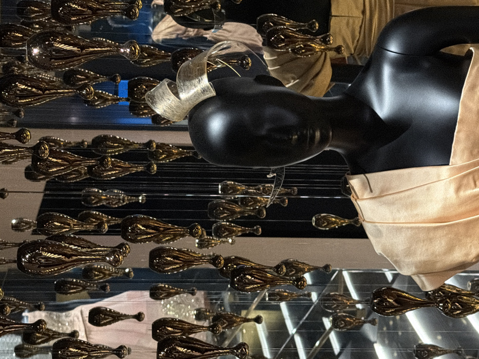 The image depicts an interior view of a luxury retail establishment, specifically an elaborate fashion display, characteristic of major Parisian department stores such as Galeries Lafayette Haussmann, located in Paris, France.
In the foreground, a glossy black, featureless mannequin torso is positioned off-center to the right, facing slightly towards the viewer. The mannequin wears a draped, light champagne-colored fabric garment, which folds over its right shoulder and across its chest. A matching, multi-looped fabric fascinator or headpiece is affixed to the mannequin's head.
The midground and background are dominated by a dense, vertical array of numerous polished, golden, teardrop-shaped objects. These objects, varying in size, are suspended from above by thin, dark vertical wires, creating a shimmering, curtain-like effect due to reflections of ambient light.
Faintly visible in the blurred background to the far left, another mannequin or display stand showcases a light-colored dress. To the right of the foreground mannequin, a partially obscured display features a garment of similar golden tone. Architectural elements, possibly stair railings or mirrored surfaces, are subtly discernible in the deepest background. The overall scene is brightly illuminated, with light reflecting prominently off the golden objects and the mannequin's surface.