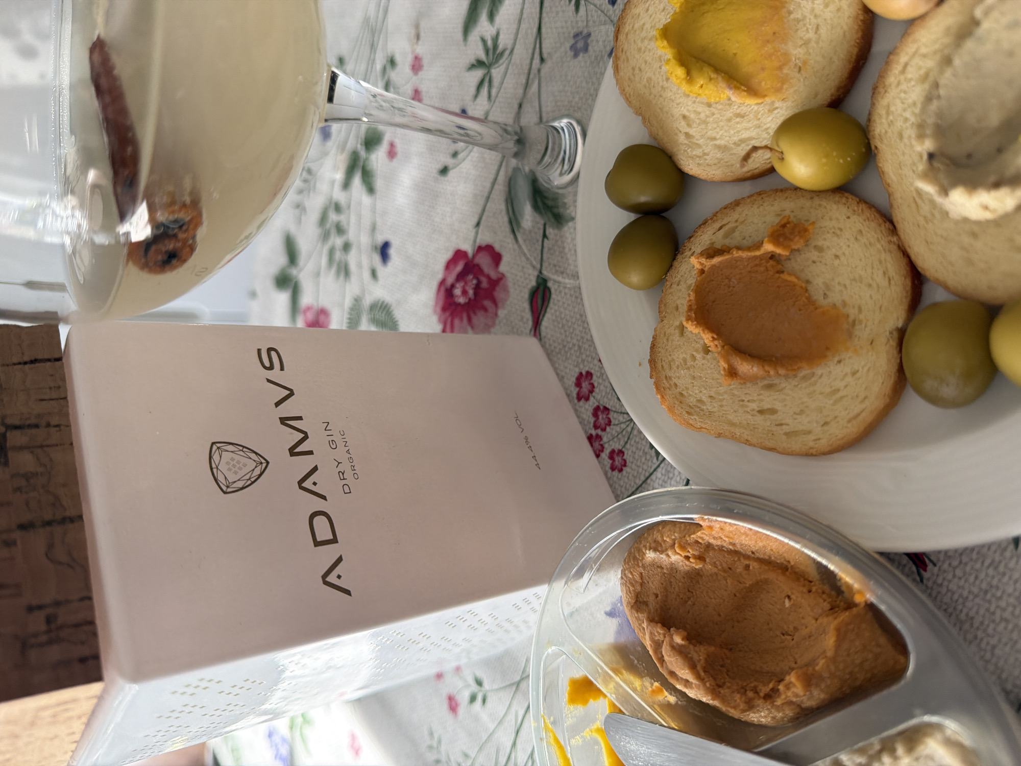 The image captures a close-up, elevated perspective of a table setting in Esch-sur-Alzette, Luxembourg. The surface is covered by a white tablecloth featuring a pattern of pink roses, green leaves, and smaller blue and purple flowers. A frosted white, rectangular bottle, identified by "ADAMVS DRY GIN ORGANIC" and "44.4% VOL" text along with a shield emblem, stands centrally. Its neck is topped with a cork stopper. To the right, a stemmed glass contains a pale yellow liquid with a single cinnamon stick submerged. In the foreground, a white circular plate holds multiple slices of toasted bread. One slice is spread with an orange-brown paste, another with a bright yellow paste, and a third (partially visible) with a light beige paste. Several whole green olives are distributed around the bread on the plate. To the left, a clear plastic partitioned container displays three distinct spreads: orange-brown, bright yellow, and light beige. A small metal knife or spreader is positioned within the orange-brown spread in the container. No individuals are visible within the frame.