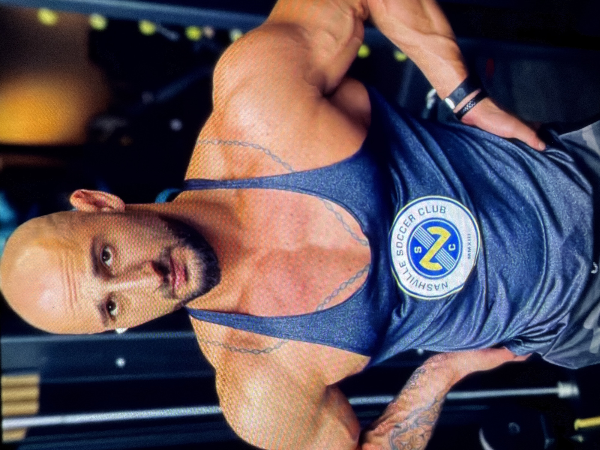A muscular, bald man with a beard wearing a blue tank top with a Nashville Soccer Club logo on the chest is facing the camera. The background features a gym setting. The exact location in Nashville, Tennessee is unknown.