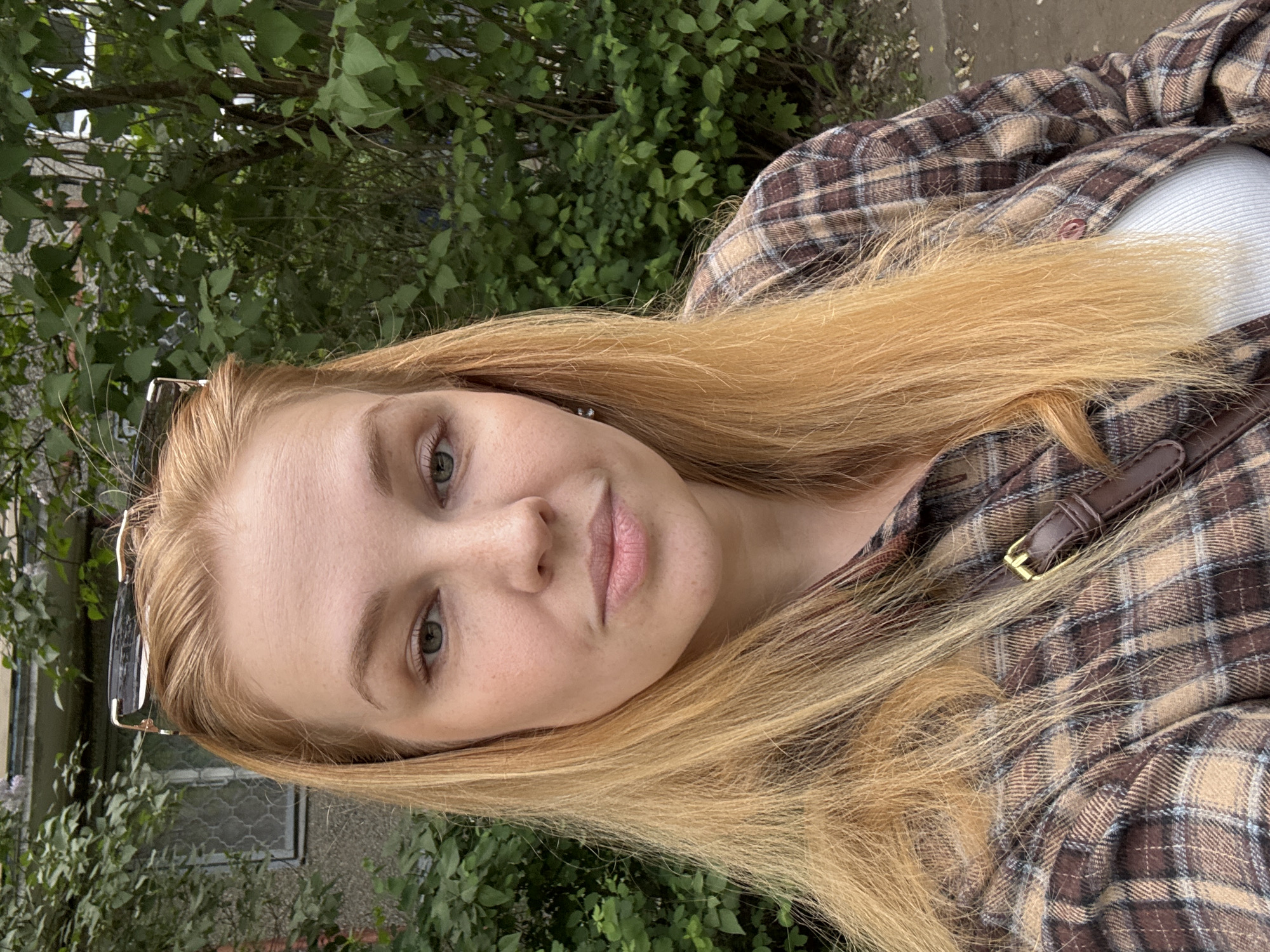 A selfie-style shot of a young woman with light complexion and long, blonde hair. She is wearing a brown and beige plaid shirt, with a glimpse of a white top underneath. Sunglasses are perched on top of her head. The background consists of green foliage, suggesting an outdoor location near vegetation. A building with a window can be seen in the blurred background. While the information provided mentions Rusanovo, Russia, nothing in the visual context confirms the location to be Rusanovo or even Russia.