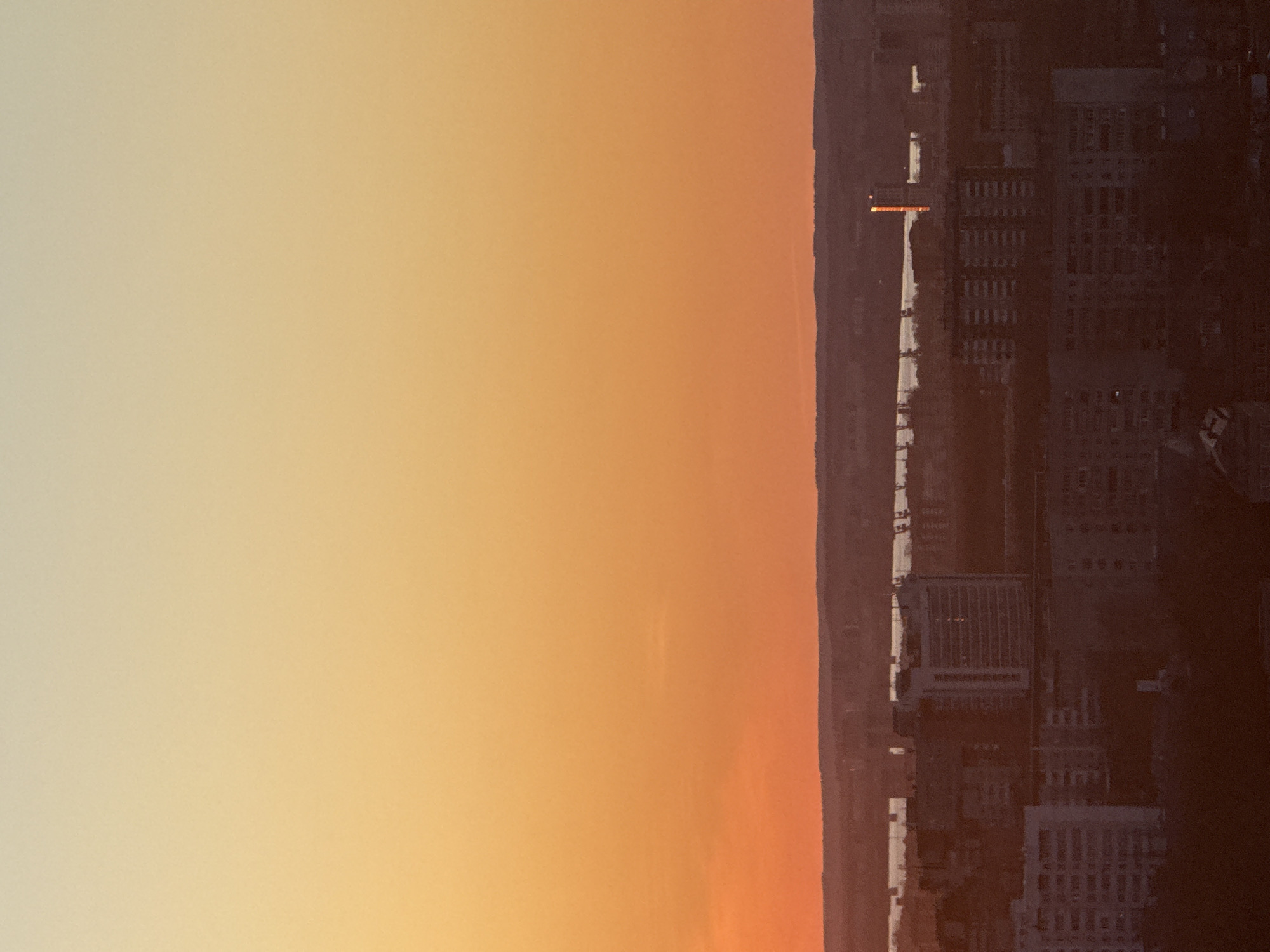The image depicts a cityscape at sunset. The sky transitions from yellow to orange. On the right side of the image are city buildings, a river, and a road. The image is a view of the city of Perm, Russia.