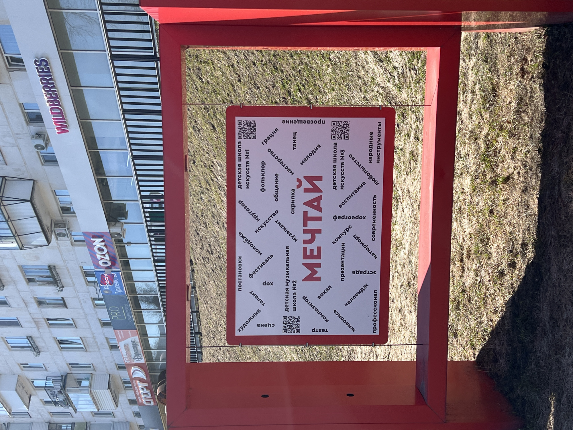 Here is a forensic description of the image:
**Scene:** The image shows a red rectangular frame structure with a sign hanging inside it. The sign is white with a red border and contains text in Russian, with the word "МЕЧТАЙ" (Dream) in large font in the center. Surrounding the word are other related words such as "art," "music," and "dance," along with QR codes.
**Setting:** The scene appears to be outdoors, with a patch of dry grass and weeds visible behind the frame. In the background, a building with multiple windows and a sign for "WILDBERRIES" is visible.
**Objects:**
* Red rectangular frame structure
* White sign with red border and Russian text
* QR codes on the sign
* Building with windows and signs in the background
* Dry grass and weeds
**Context:** The presence of the Russian text, the commercial signs, and the architectural style suggest that the location is likely in Russia.
**Geographic Context:** The building signs contain Russian text as well as the name “WILDBERRIES” indicating that it is in Russia. The sign itself reads “Детская школа искусств №1” and “Детская школа искусств №3”, which translates to “Children’s School of Arts No. 1” and “Children’s School of Arts No. 3”. A search of these translated names, in conjunction with the signs, led to the conclusion that this photo was taken in Chaikovsky, Russia.