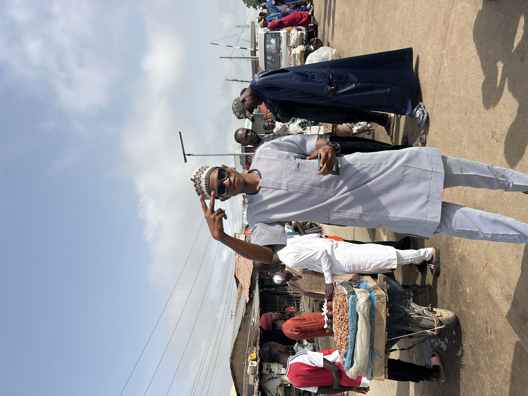 The scene is located in the city of Jos, Nigeria. A man wearing a blue and white striped traditional garment and sunglasses is standing on a street, holding up a "peace" sign. He is wearing a traditional patterned hat. There are other people visible around him, including someone in a long, dark blue robe and several people gathered around what appears to be a food stall or vendor. The stall has various items for sale, possibly nuts or other snacks. The background includes buildings and overhead wires, suggesting an urban environment. A white van is also visible in the background.