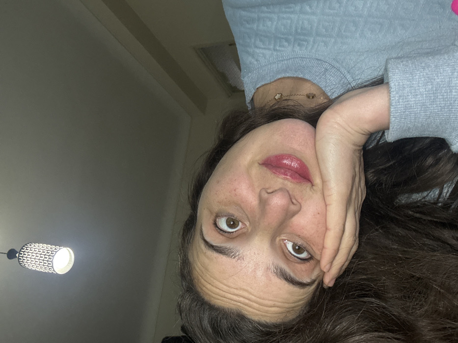 A woman with brown hair and eyes poses upside down. Her face is partially obscured by her hand, which rests against her cheek. She wears a light blue sweater and a delicate gold necklace. Her lips are painted with a glossy, pink hue. The background features a dimly lit room with a ceiling light fixture visible in the lower left corner. The lighting is soft, accentuating the wrinkles on her forehead.